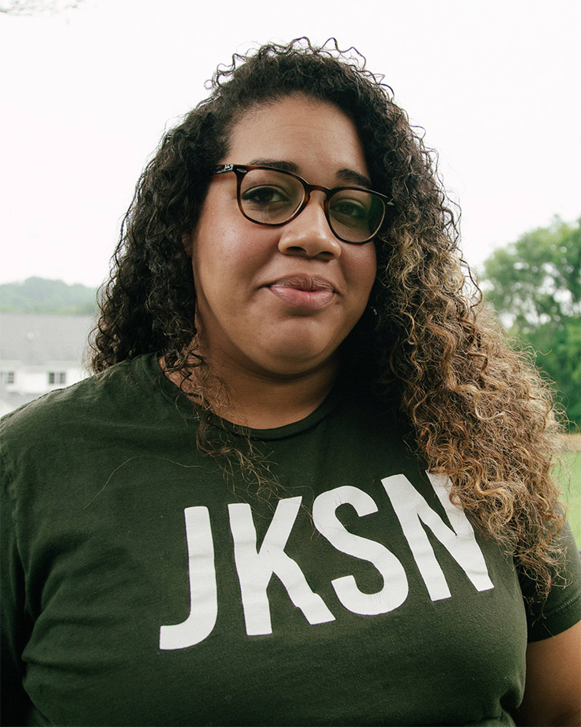 A woman with medium complexion, curly brown hair and glasses. She has a black Tshirt with the letters "J K S N" on it.
