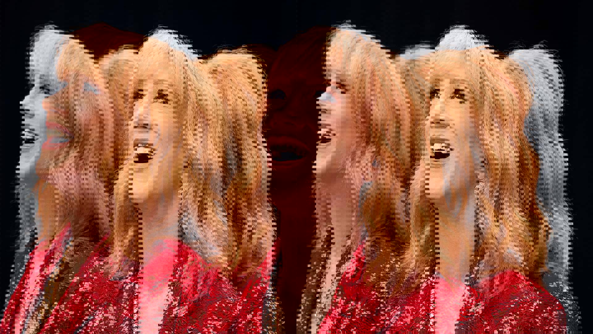 A smiling middle aged woman with light complexion, medium length wavy red hair, and blue eyes. and a red top. She is standing against mirrors such that her reflection is showing three other times in different directions. 