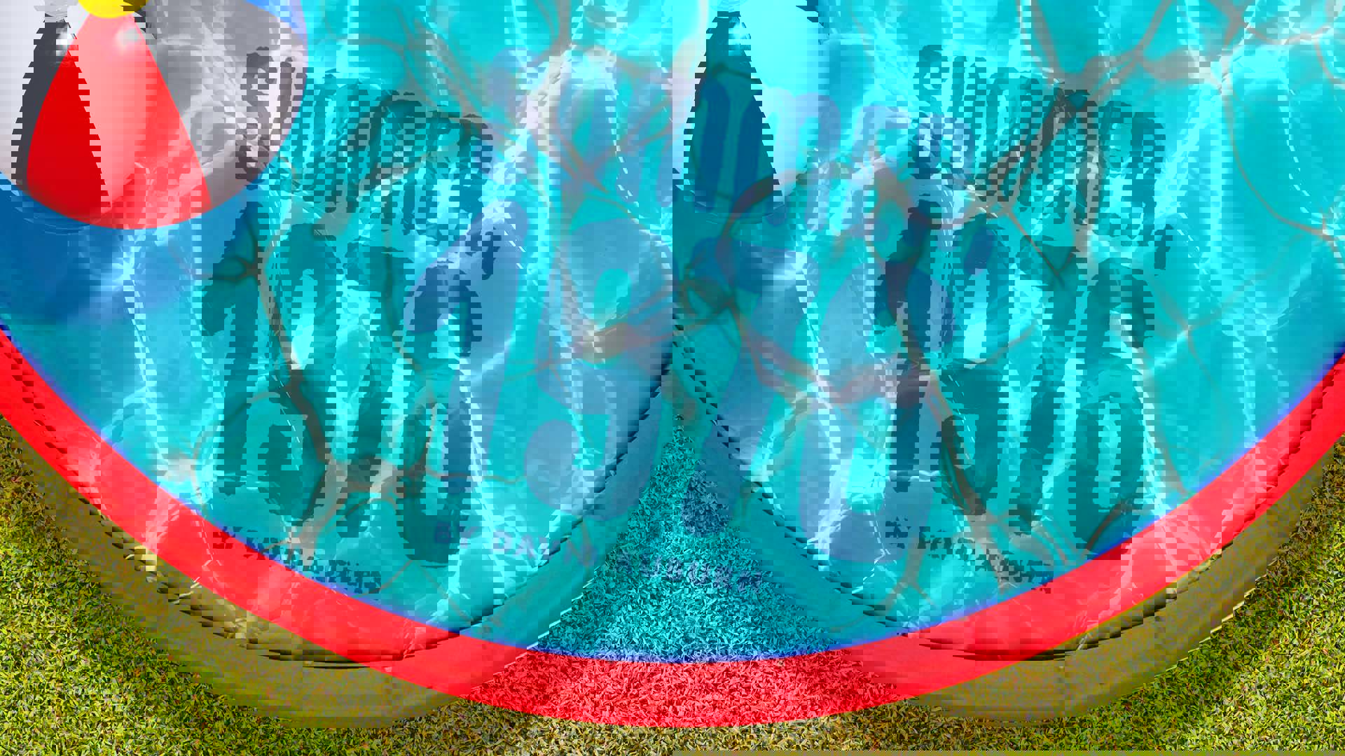 A small, red, blow-up plastic swimming pool filled with light-reflecting vibrant blue water with grass below, large blue text in the water reads: "Summer, 1976 by David Auburn.