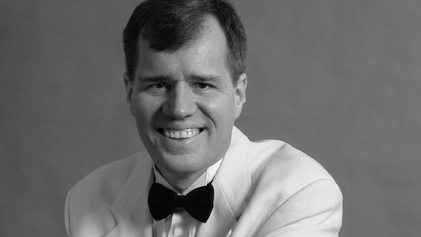 Greyscale photo of a a white man with short hair and dark eyes in a white tuxedo with a handkerchief leaning against a piano.