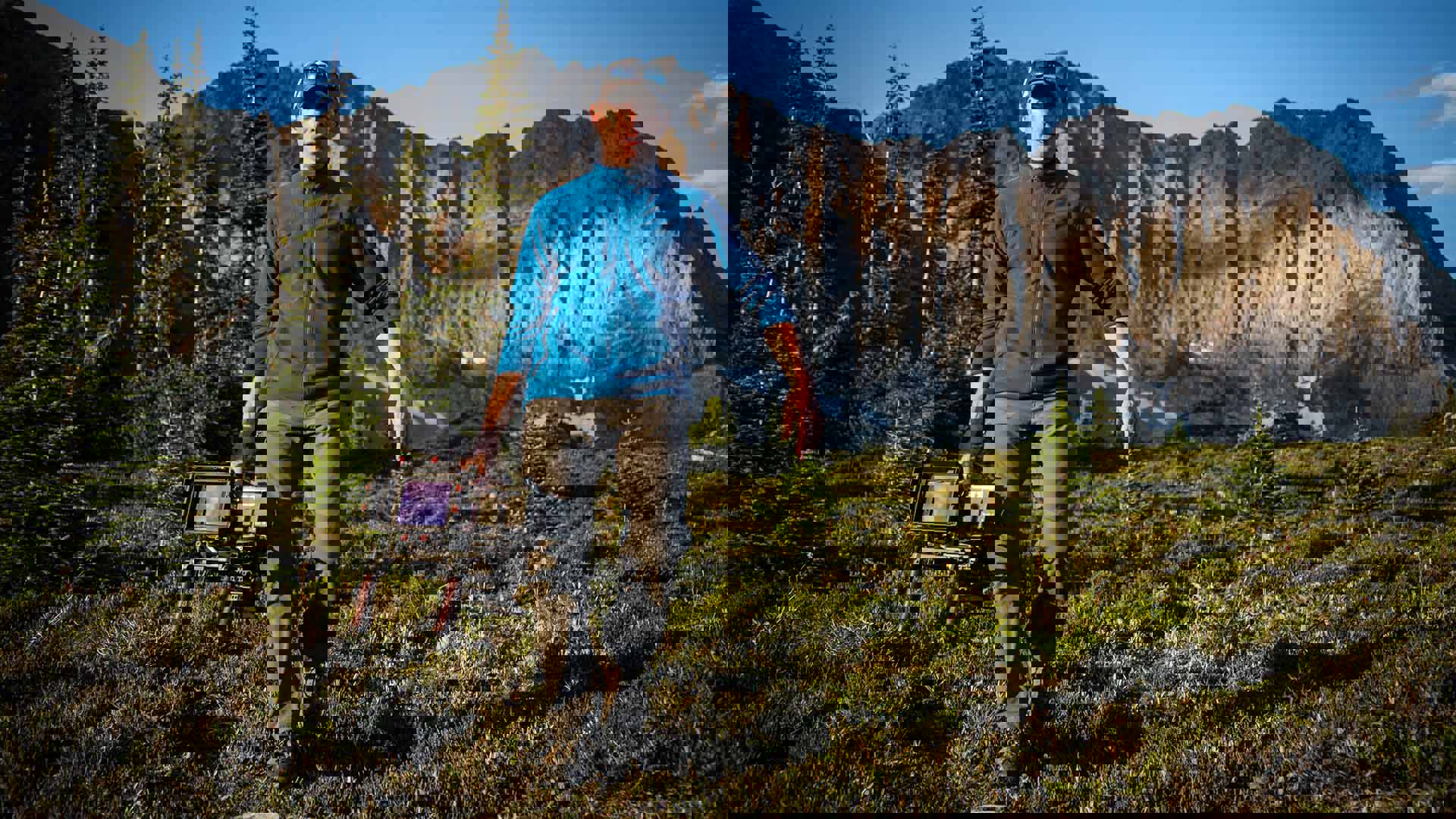 A caucasian man holding a video camera walking down a grassy hill backed by mountainous terrain.