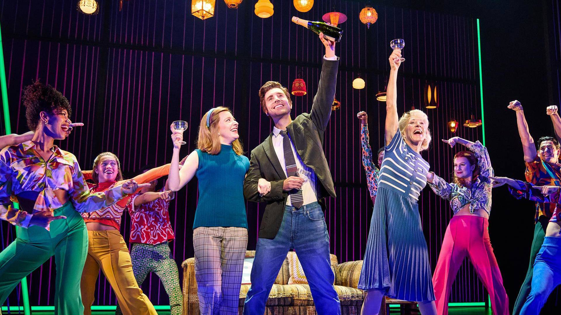 A man with light complexion and short brown hair in a grey jacket and jeans holding a champagne bottle above his head and hooking arms with a woman with light complexion, medium reddish hair and a blue top. They are surrounded by people dancing in colorful clothes.