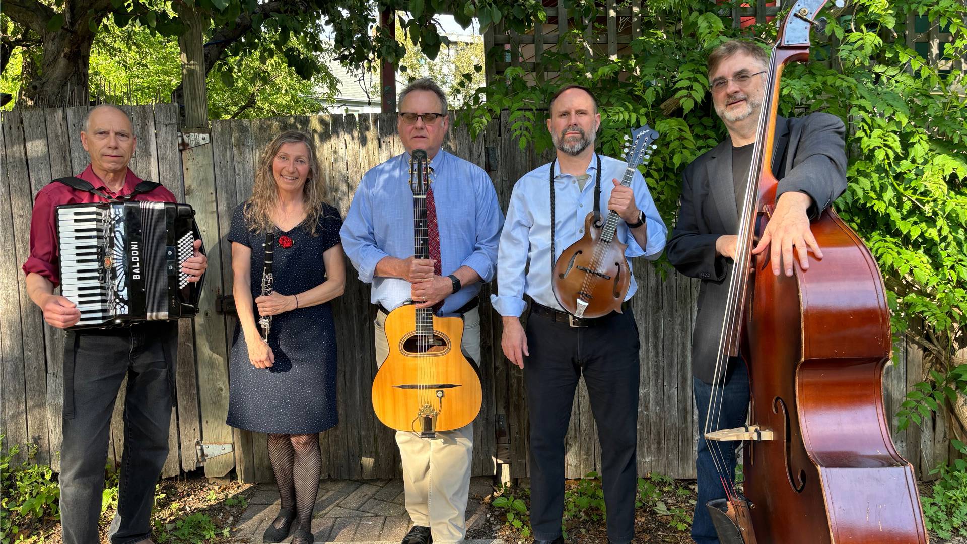 Five middle aged musicians stand outside in front of a fence holding their instruments. 