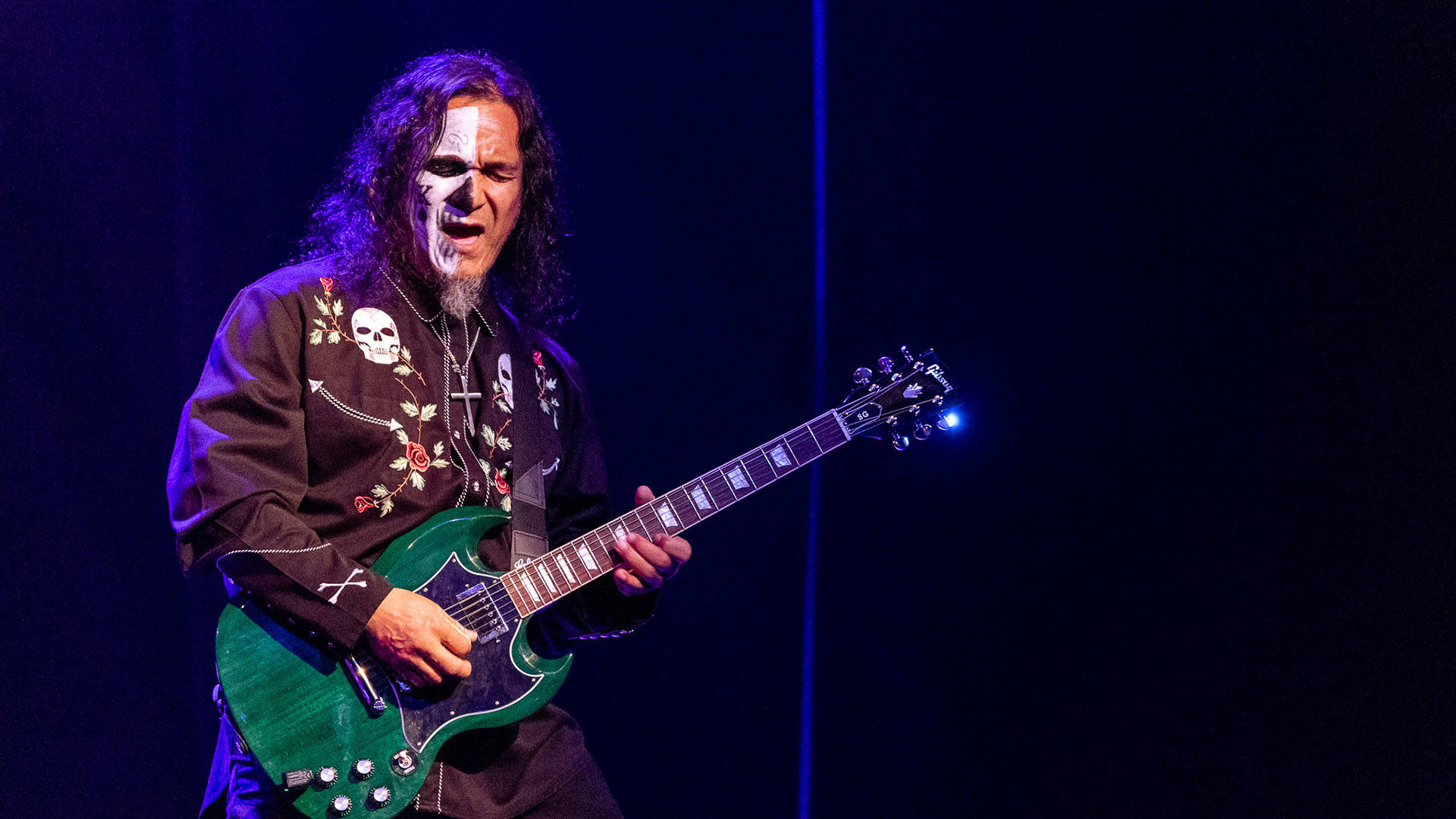 A man with dark hair wearing a shirt with flowers and skulls plays a green electric guitar.