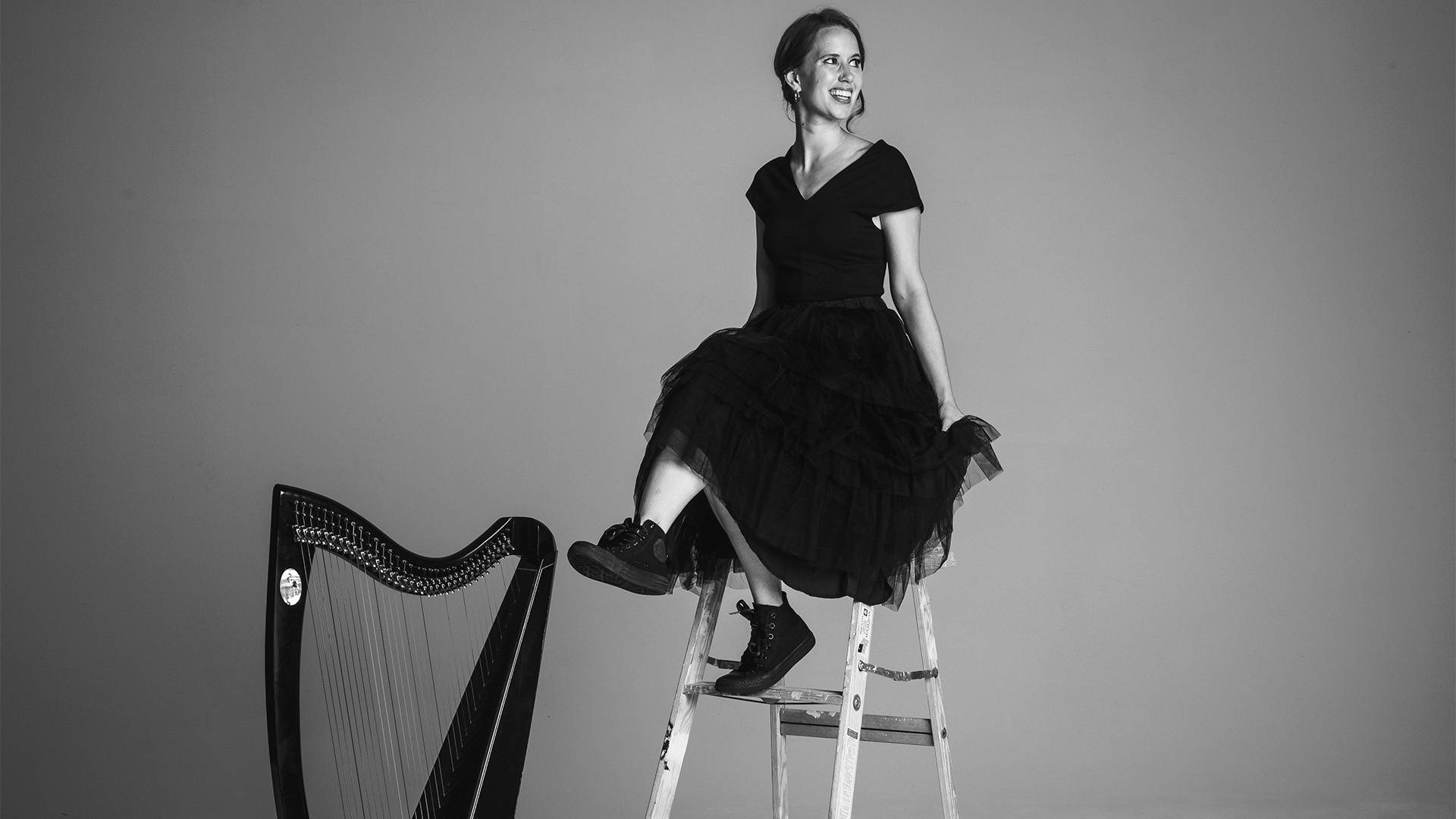 Greyscale image of a white woman with dark hair and a dark dress sitting on a ladder smiling and looking to her left. A harp is on the ground to her left.