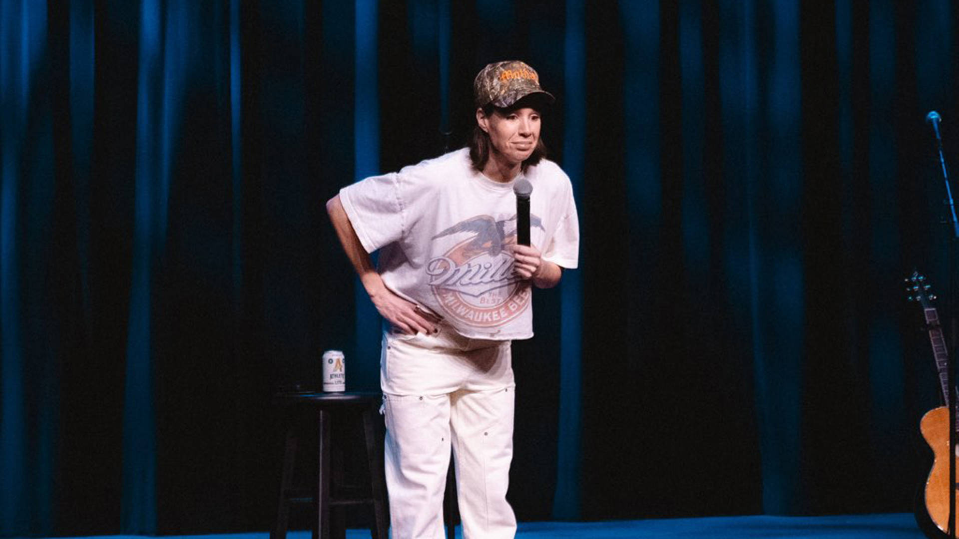 Danae Hays, a woman with light complexion, medium length brown hair, a hunter camo baseball hat and a baggy Miller brand tshirt, performing standup.