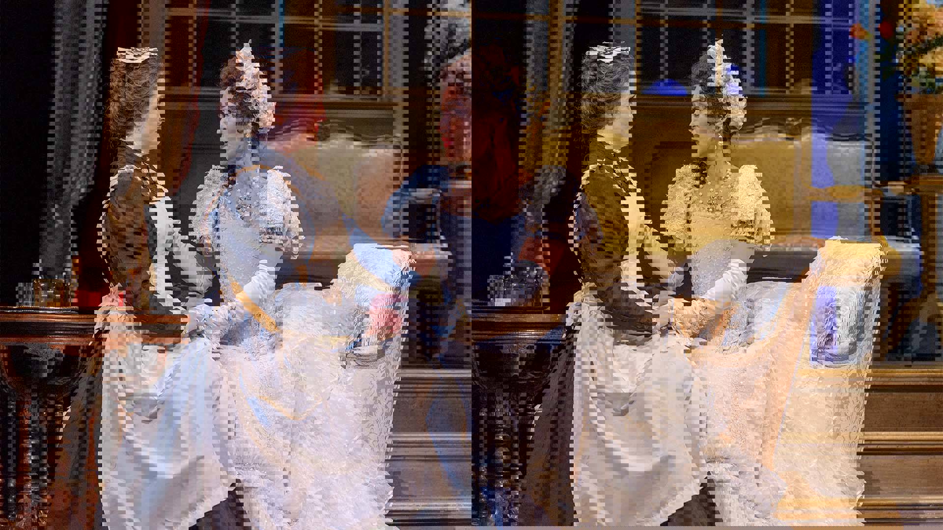 Two women performing in an opera. One is wearing a maid's outfit and the other an elegant dress. They are sitting and holding hands.