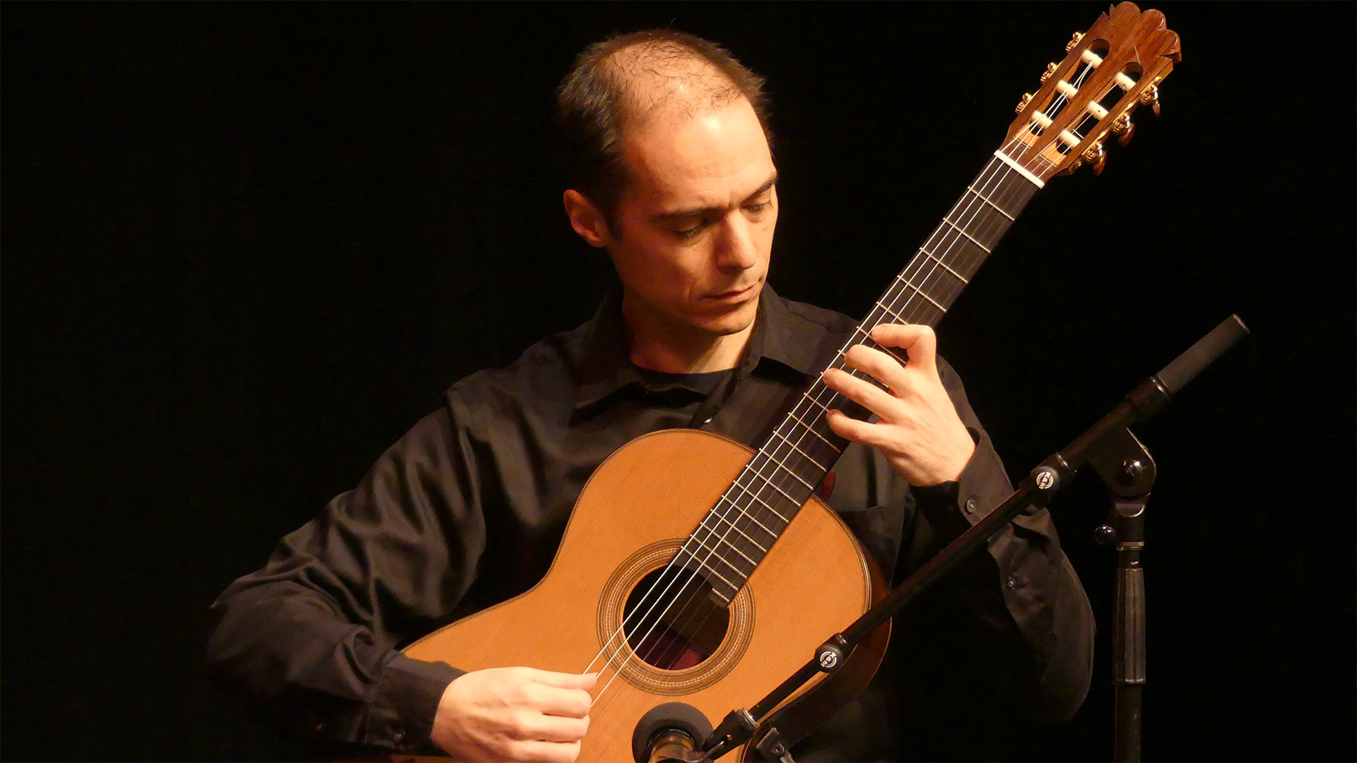 A man with medium complexion, a balding head with dark hair and black clothing playing a classical guitar.