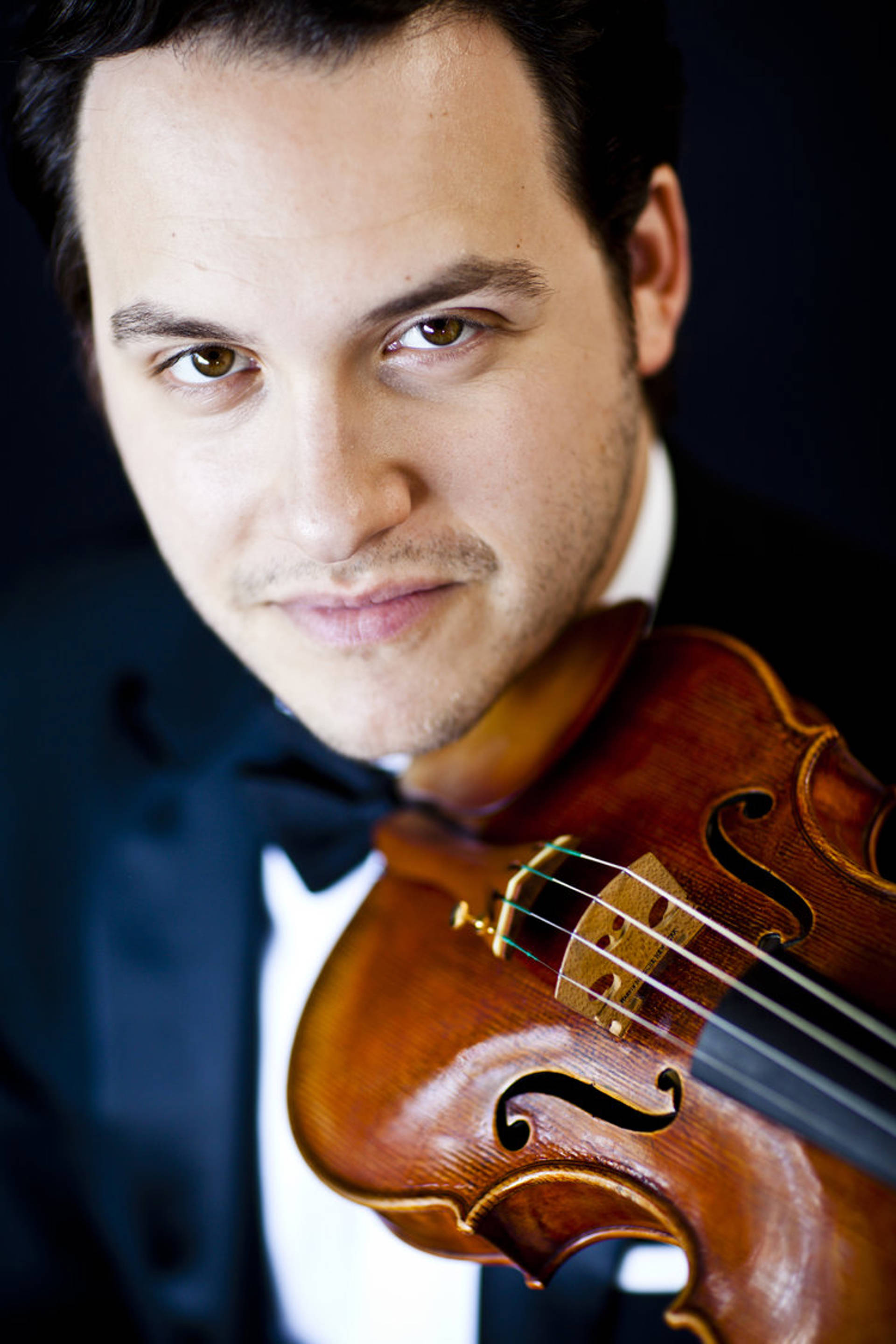 Headshot of a young man with light complexion, dark hair and brown eyes. He is holding a violin.