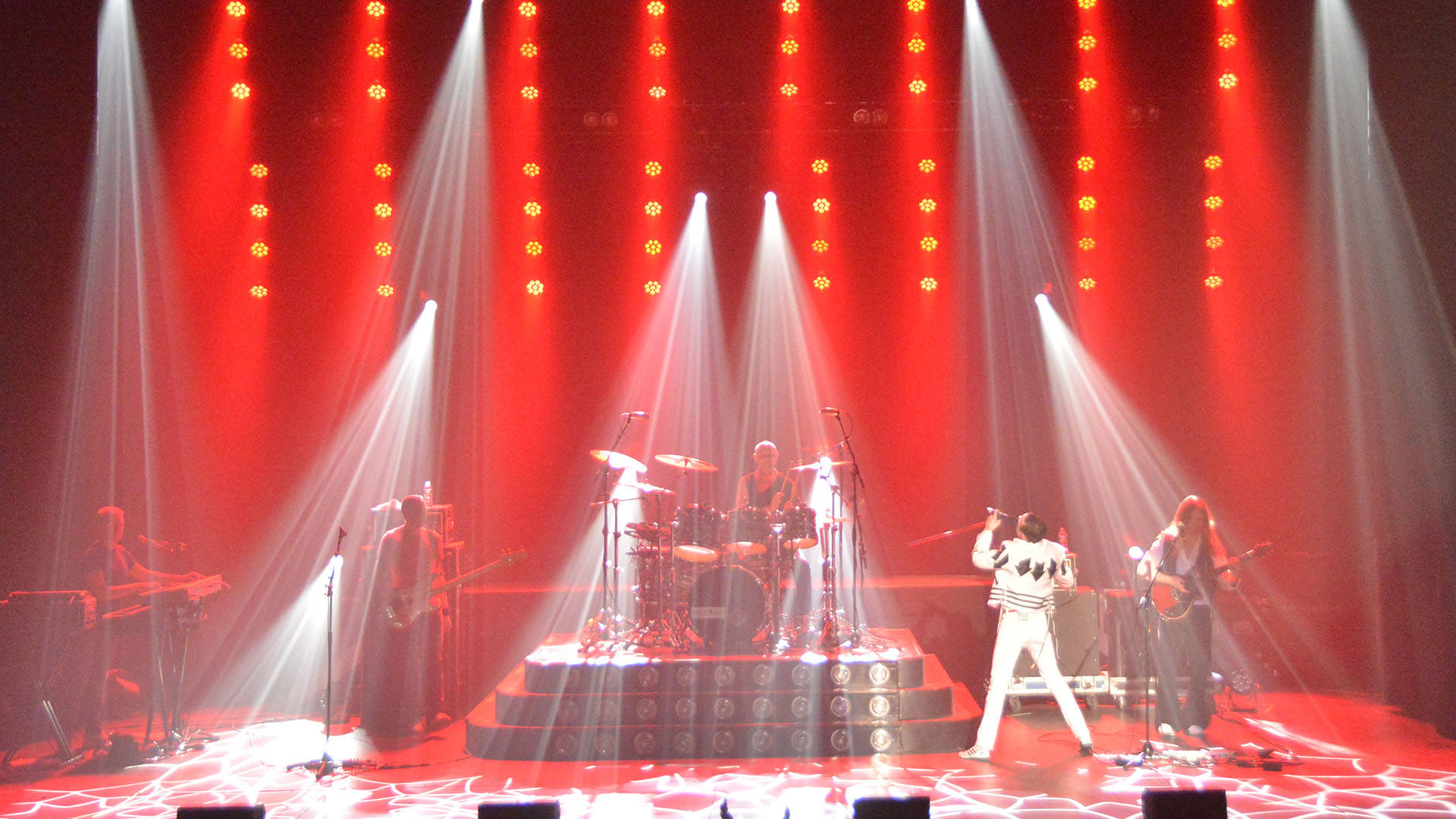 A Queen cover band featuring a singer in a white outfit impersonating Freddie Mercury. The stage is cast in red lights
