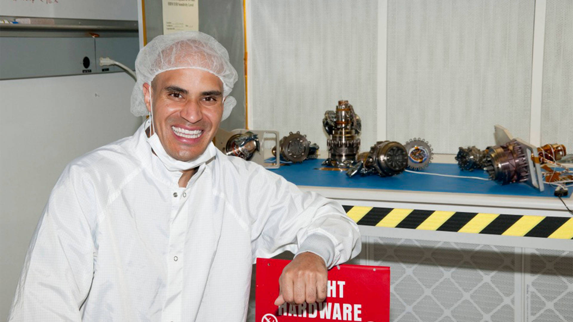 Kobie Boykins, a Black man wearing a white lab coat, mask and hair net standing next to various mechanical components.