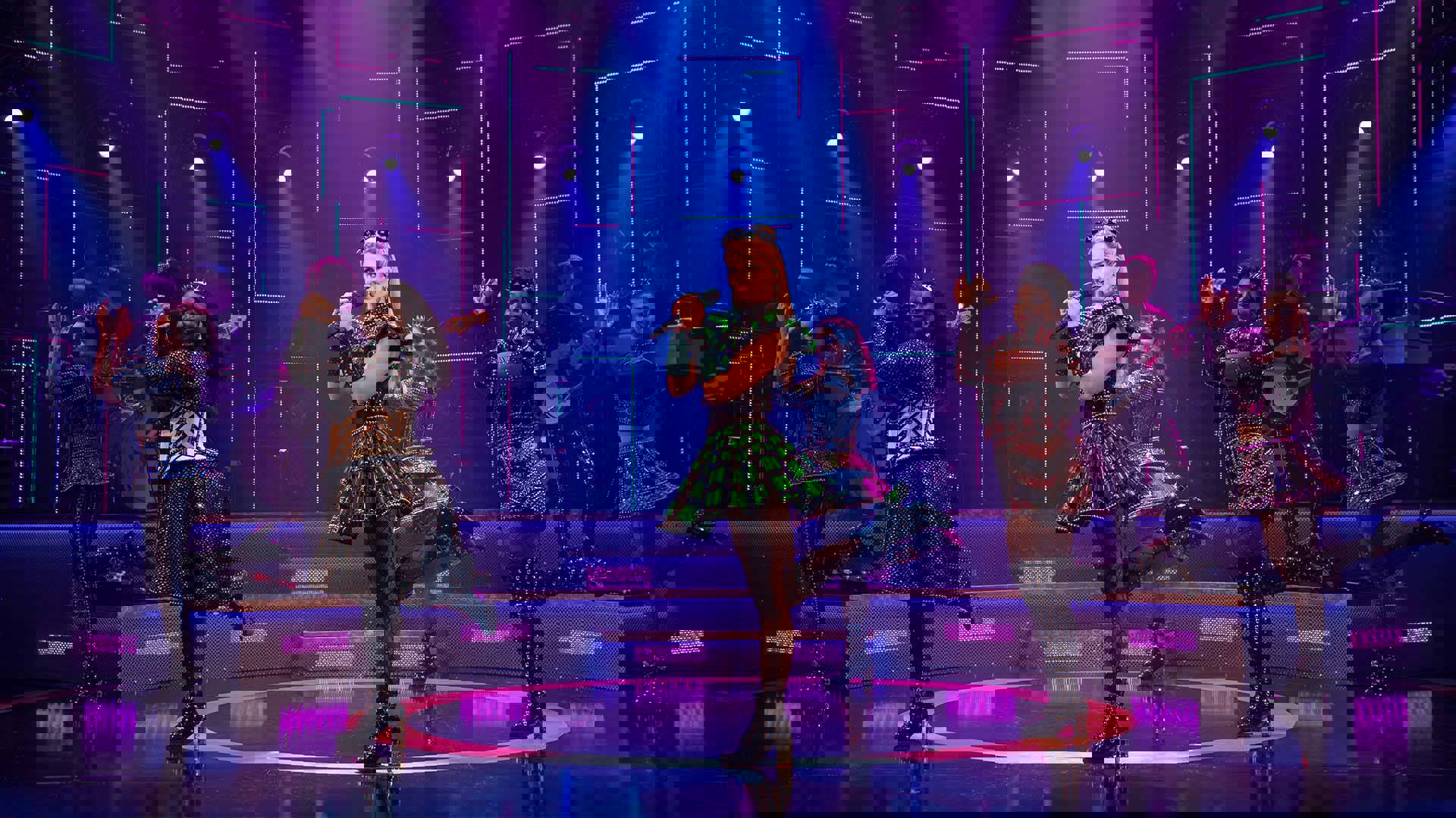 The cast of Six, in futuristic glittery outfits and holding microphones, performing on stage