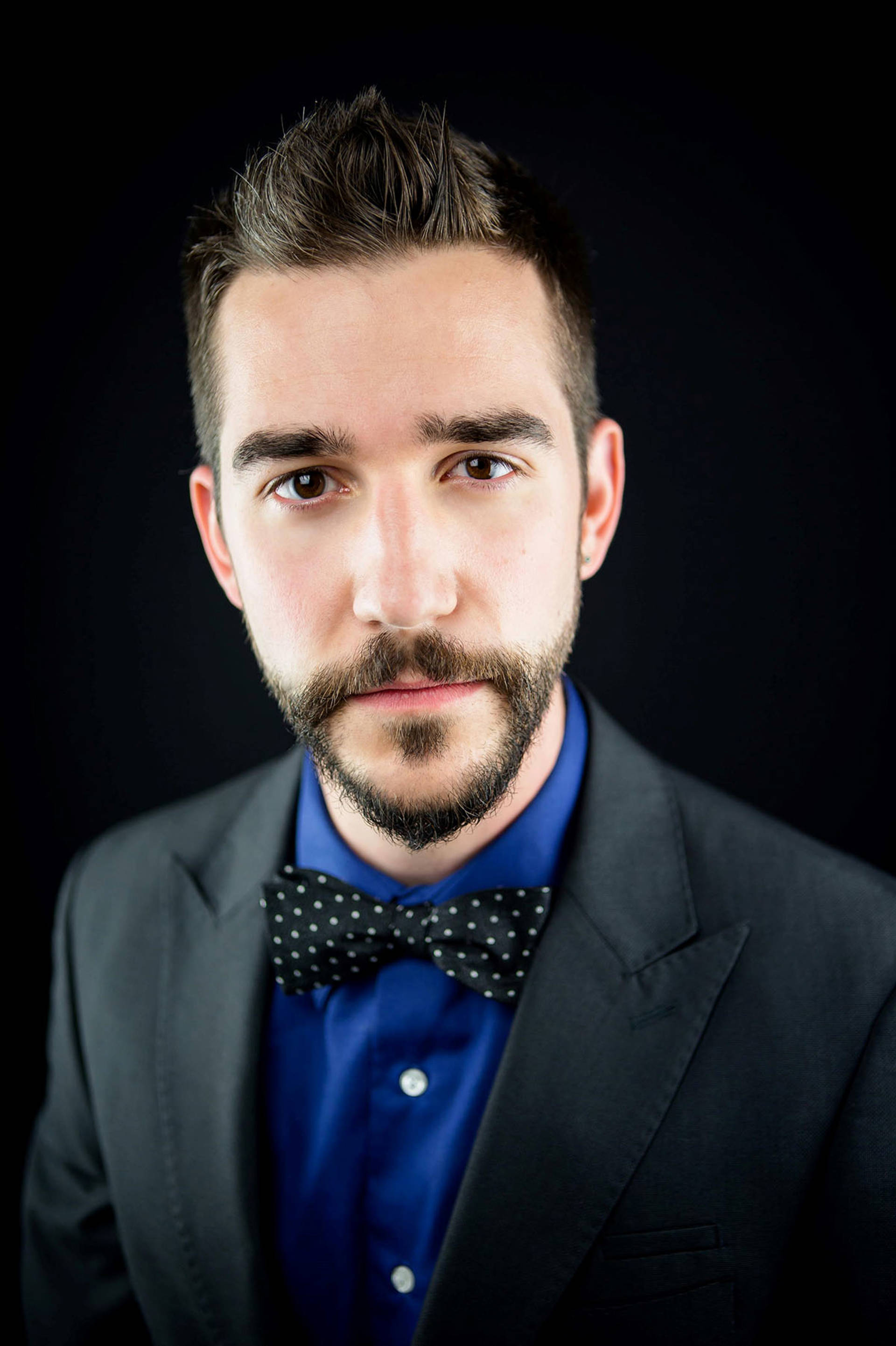 Headshot of a caucasian woman with short brown hair , dark eyes, a beard, and suit with black bowtie.