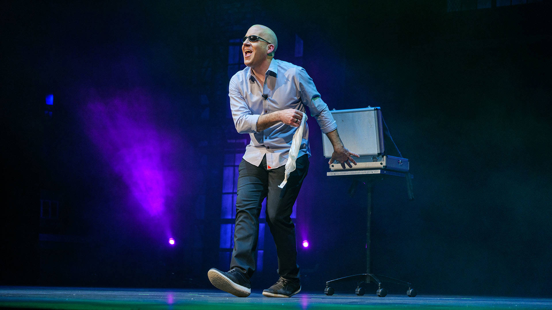 A white bald man wearing sunglasses on stage, wearing a blue button down shirt and dark jeans.