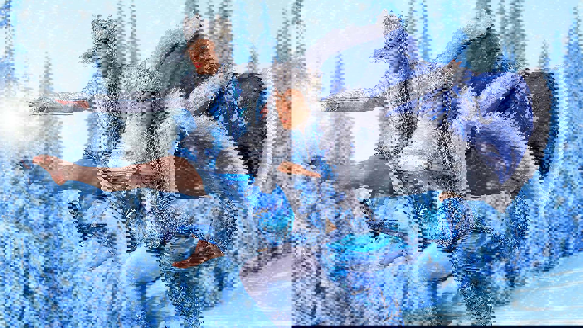 Two dancers with light complexion are leaping in ornate winter-ice themed white & blue fur-trimmed costume and bejeweled crowns against a snowy winter forest.