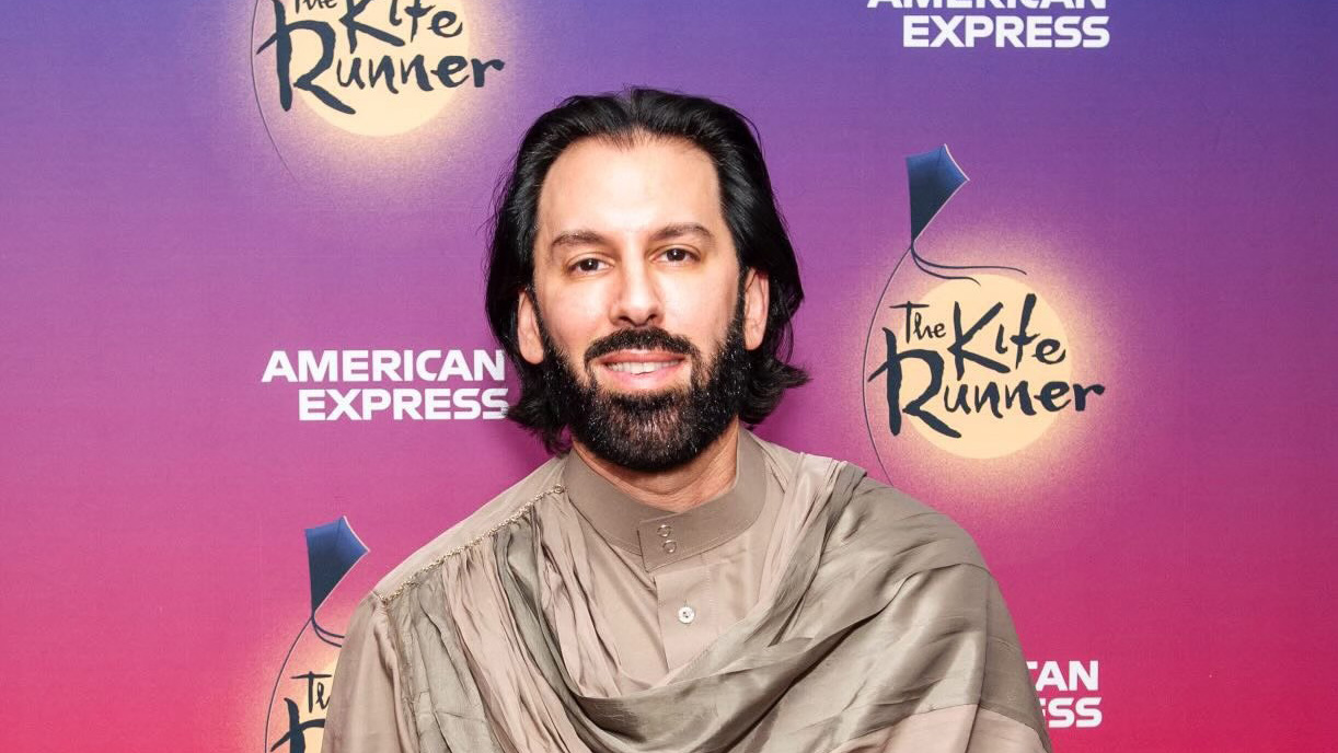 A man with medium complexion, medium dark hair and a dark beard stands and smiles. He is wearing a tan outfit.