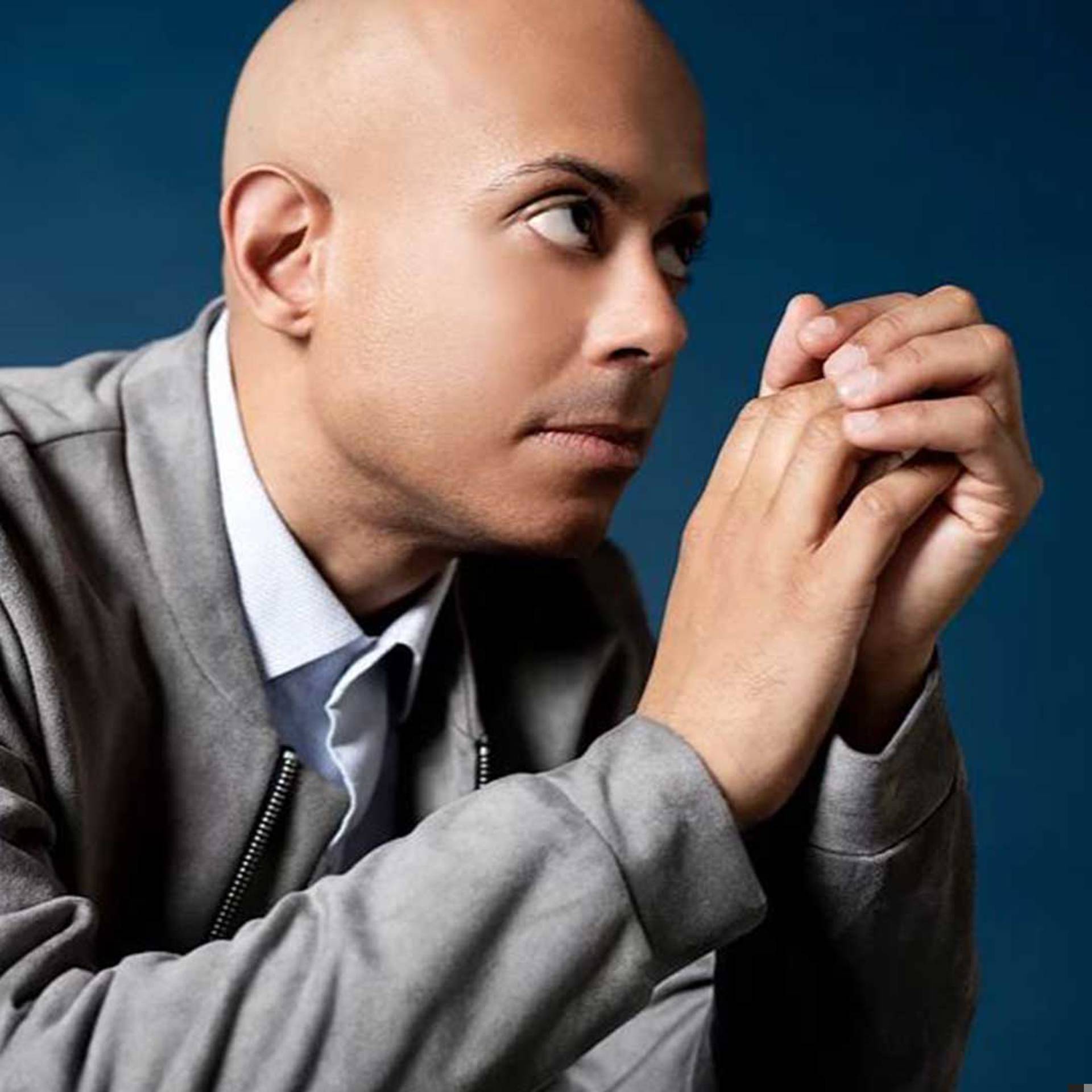 A young african american man with with a bald head is looking sideways and holding his hands together in front of his face. He has a light blue collared shirt on and a grey velvet jacket.