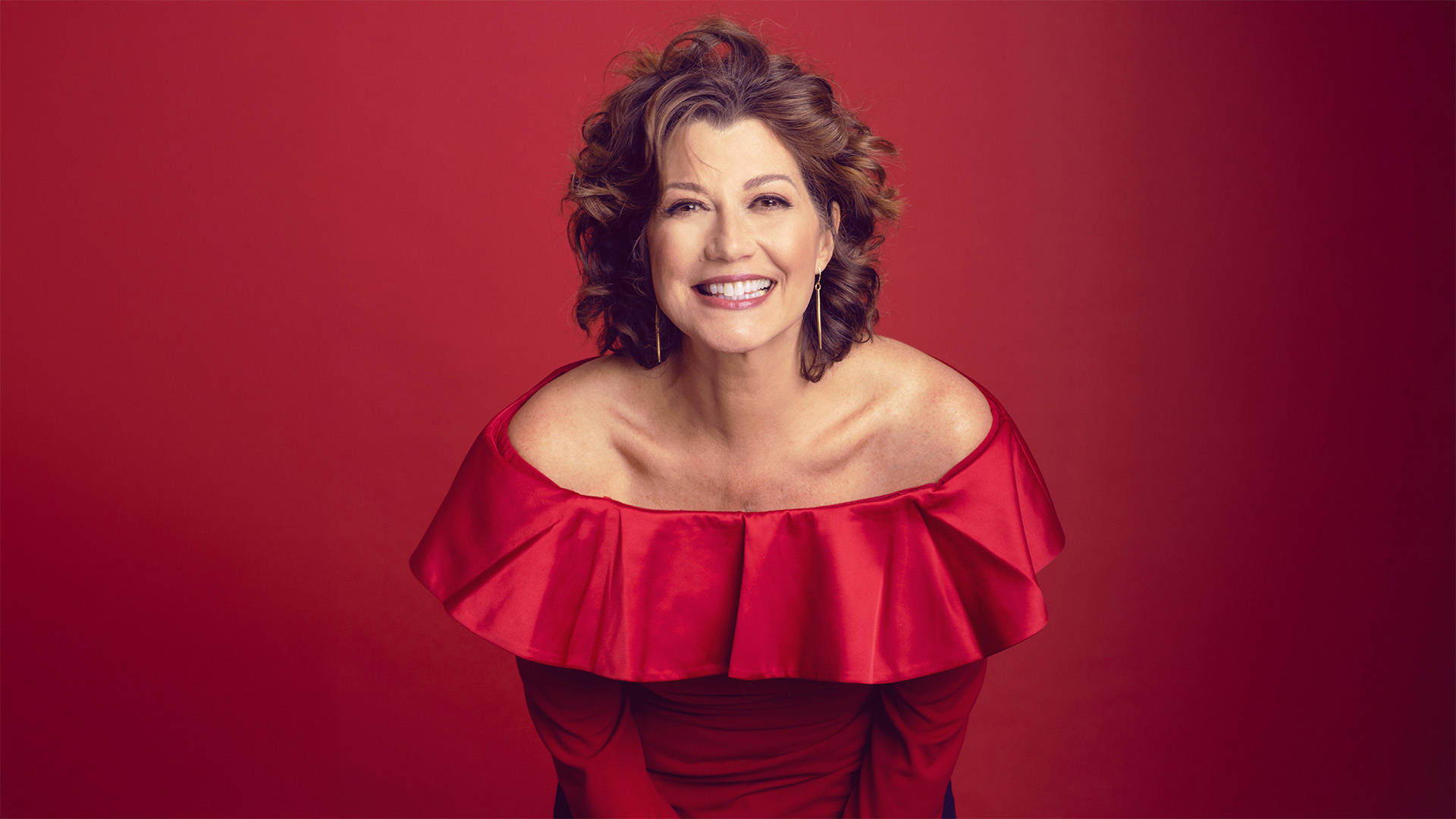 A middle-aged caucasian woman with wavy brown medium length hair. She is smiling and wearing gold earrings and a red silk wide neck blouse against a dark red background.