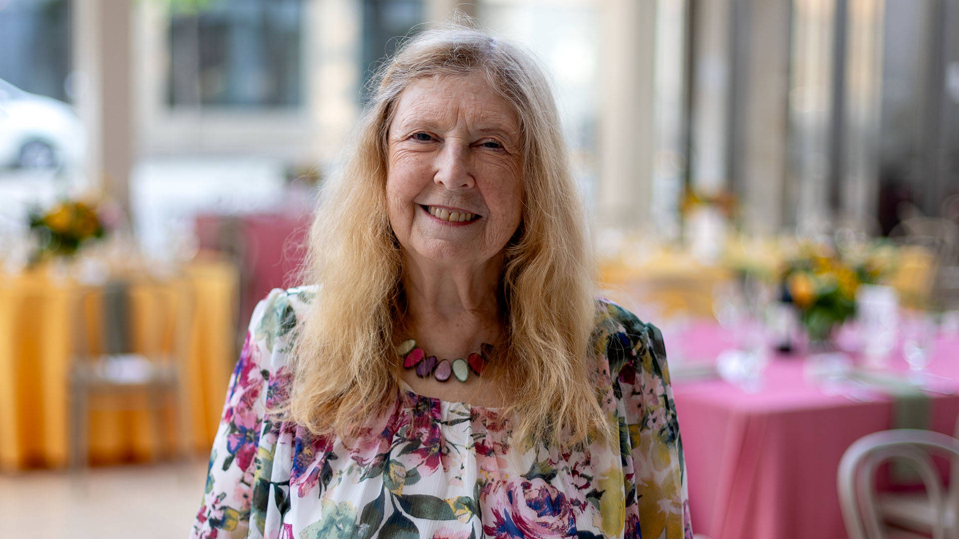 An older, caucasian woman with long blonde hair smiling in a white and pink floral dress with a matching stone necklace.