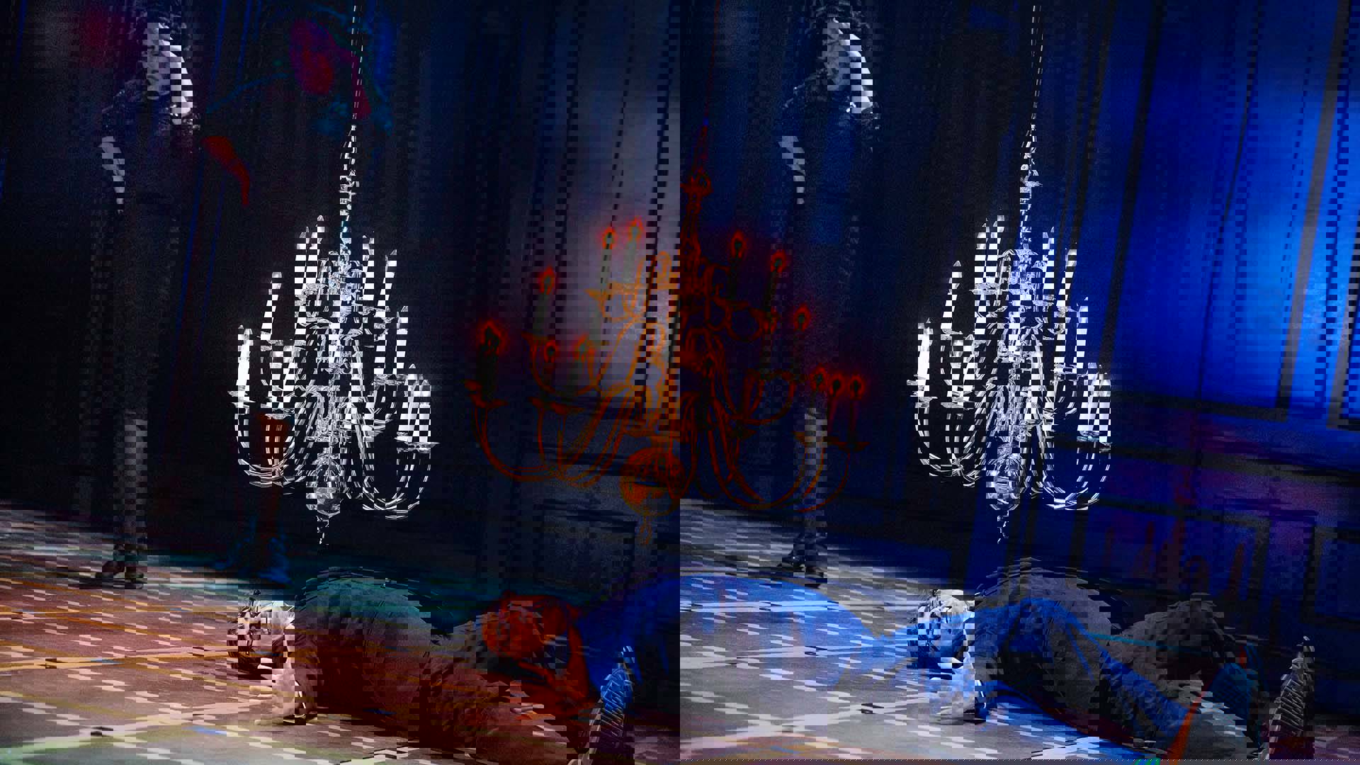 A man is lying spread out on the floor, mouth agape as a falling chandelier hangs over him. A woman in all black stands in the foreground with her hand covering her eyes.
