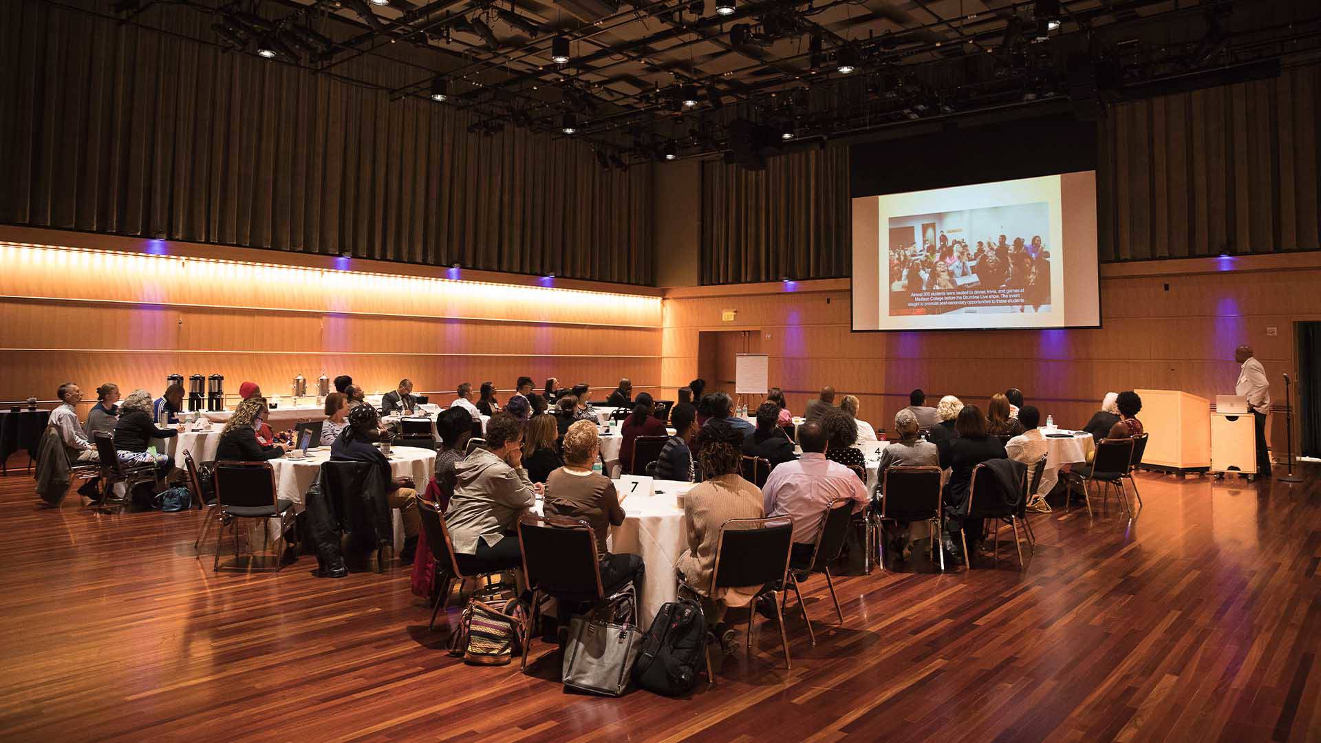 Many people sit at circular tables watching a presentation on a projector.