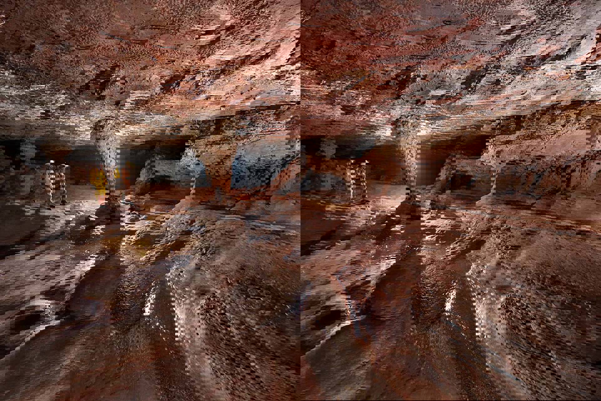 Two people in yellow and orange outfits walk through a vast rust colored cave