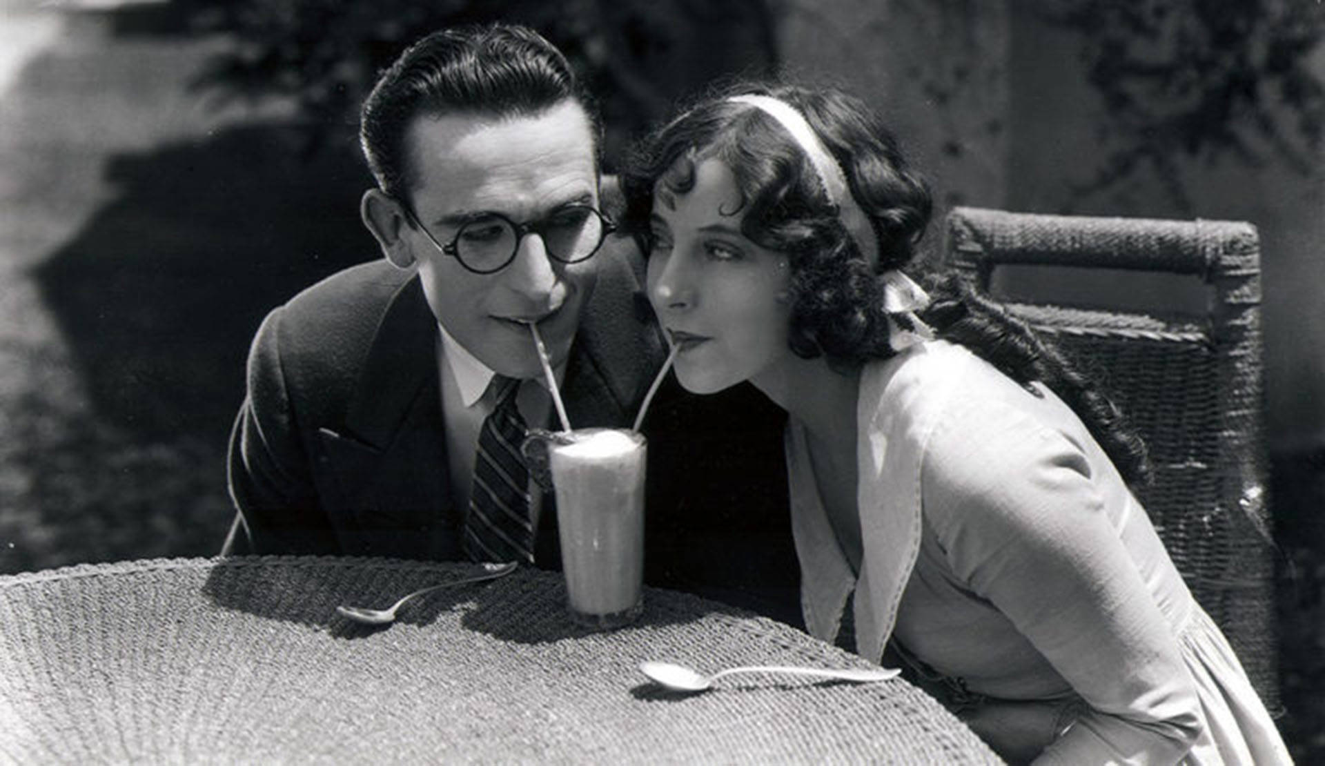 Light complexion man and woman sip on a milkshake using two straws in a scene from "For Heaven's Sake (1926)."