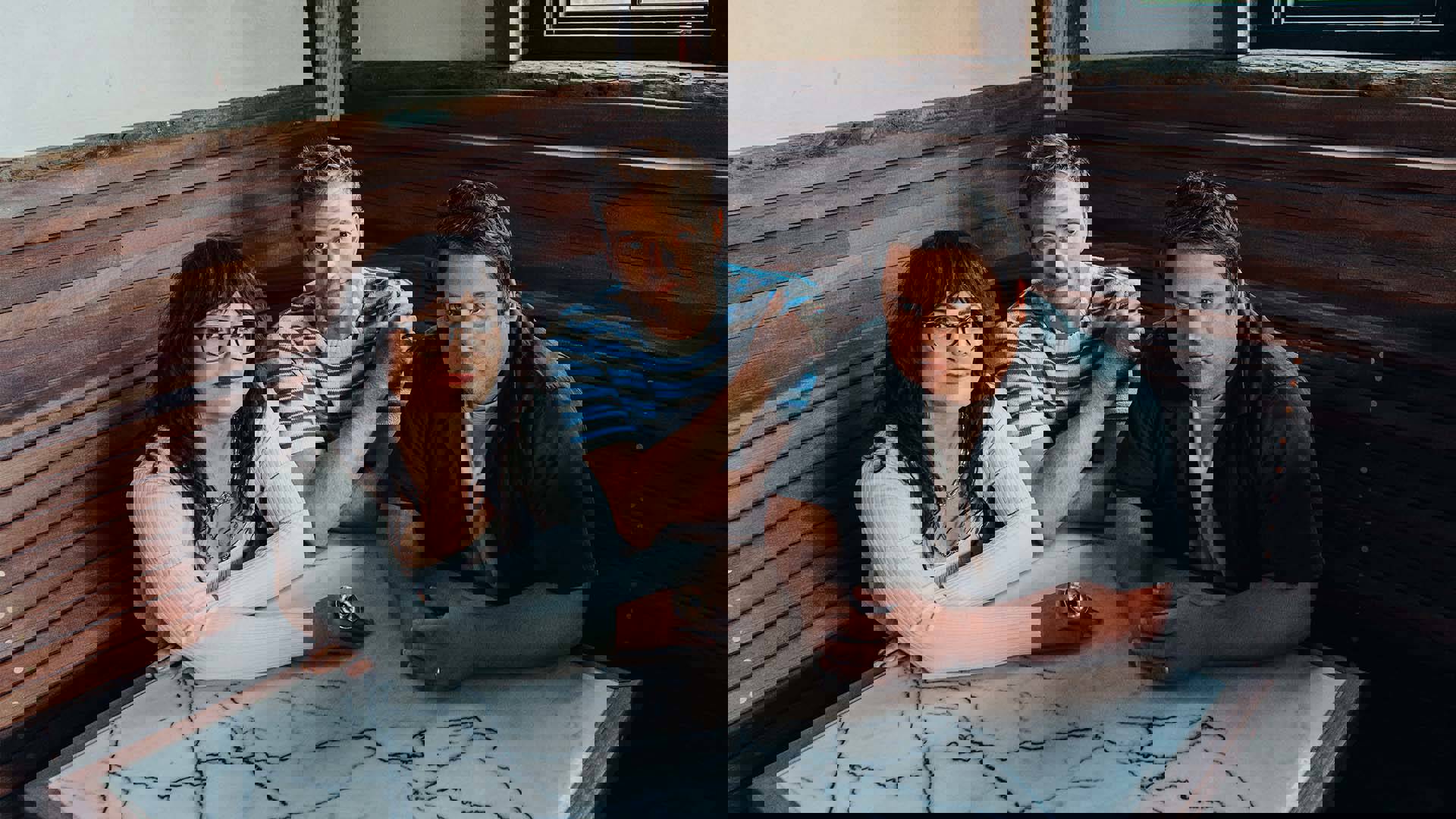 Three people with light complexion sit at a granite-design table in a corner of a room. The woman on the left has long brown hair and a white longsleeve top. The man in the middle has short blond hair and a striped blue and white sweater. The man on the right has short brown hair and an open blue buttondown shirt over white tshirt.