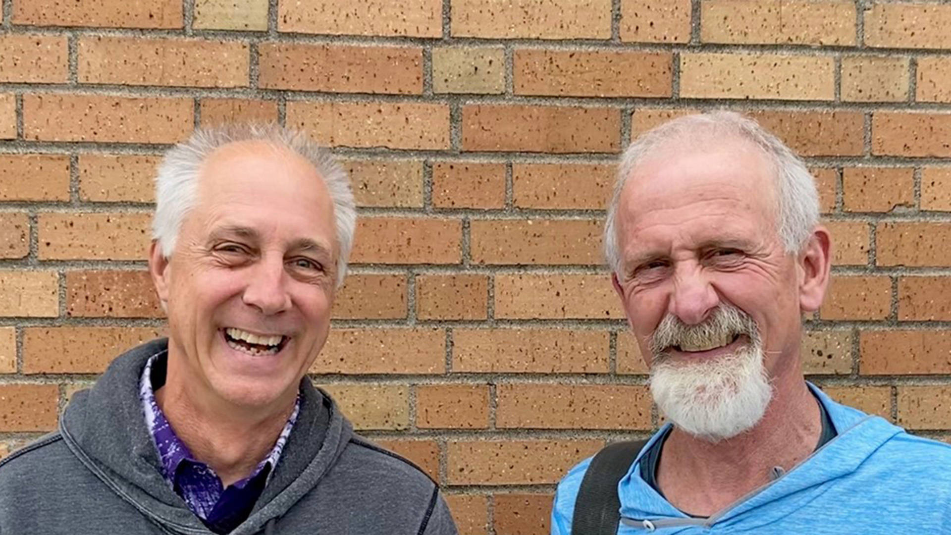 Two older white men with grey hair smiling against a brick wall.