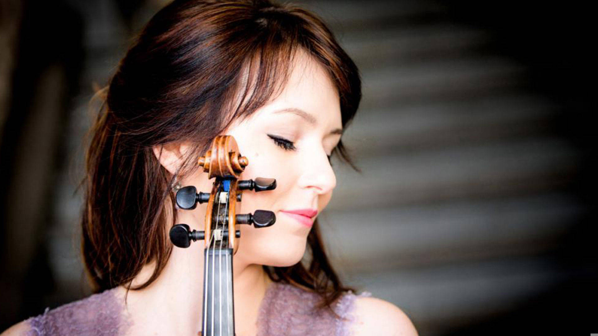 A young adult caucasian woman with dark hair with red highlights turns her head to the side and closes her eyes. Part of a violin is in the frame.
