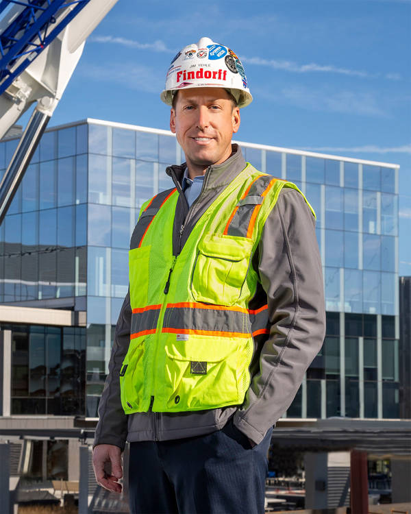 Headshot of Jim Yehle, a white man with short brown hair and blue eyes, wearing a construction hat, neon vest and grey jacket. A building with glass windows is in the background.