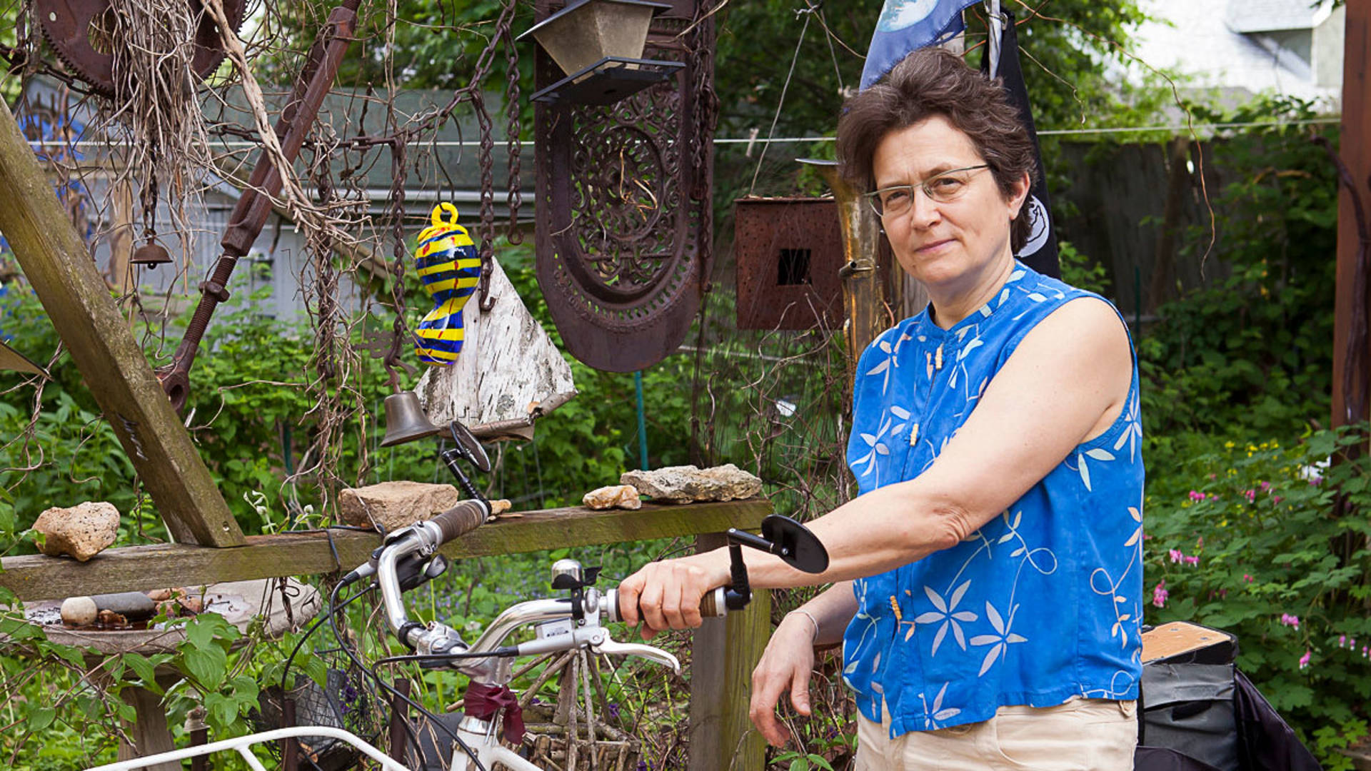 A caucasian woman with short brown hair, glasses, a blue top poses for a photo. Her left hand is resting on the handlebar of a bike.