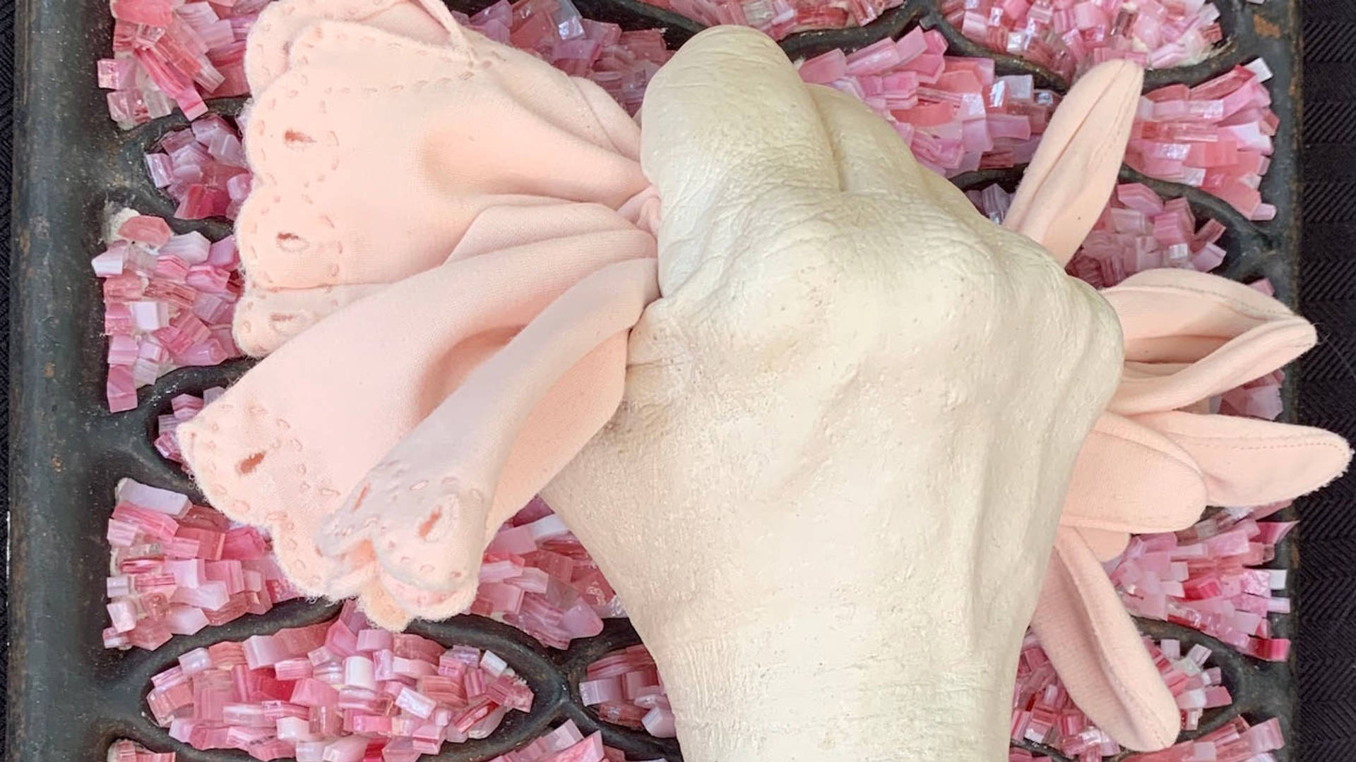 A painted white fist gripping two pastel pink gloves against a pink glass and dark metal background.