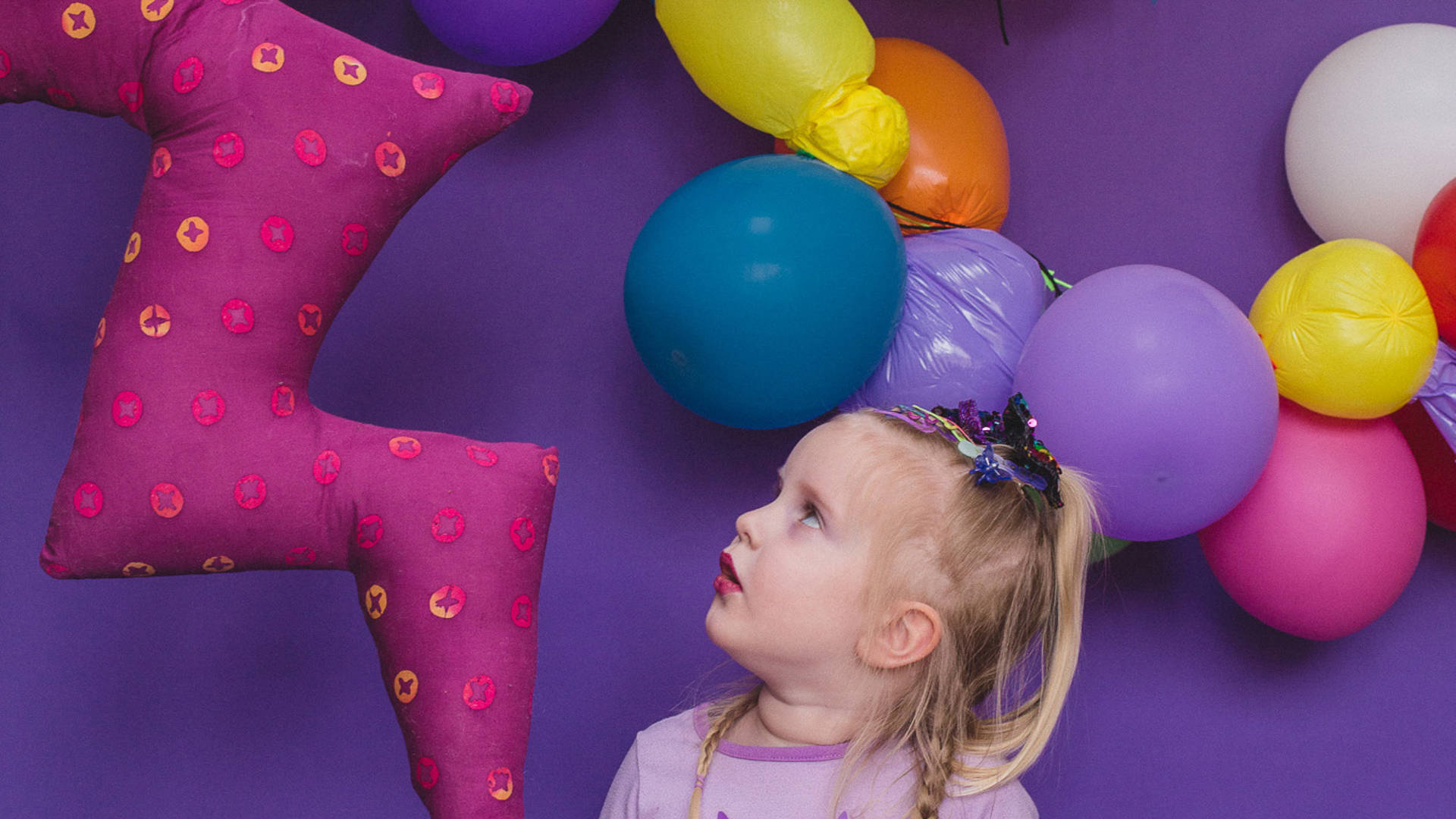 A young blond caucasian child with a purple glittery bow in their braided hair looking up at a fabric and balloon display. Purple background.