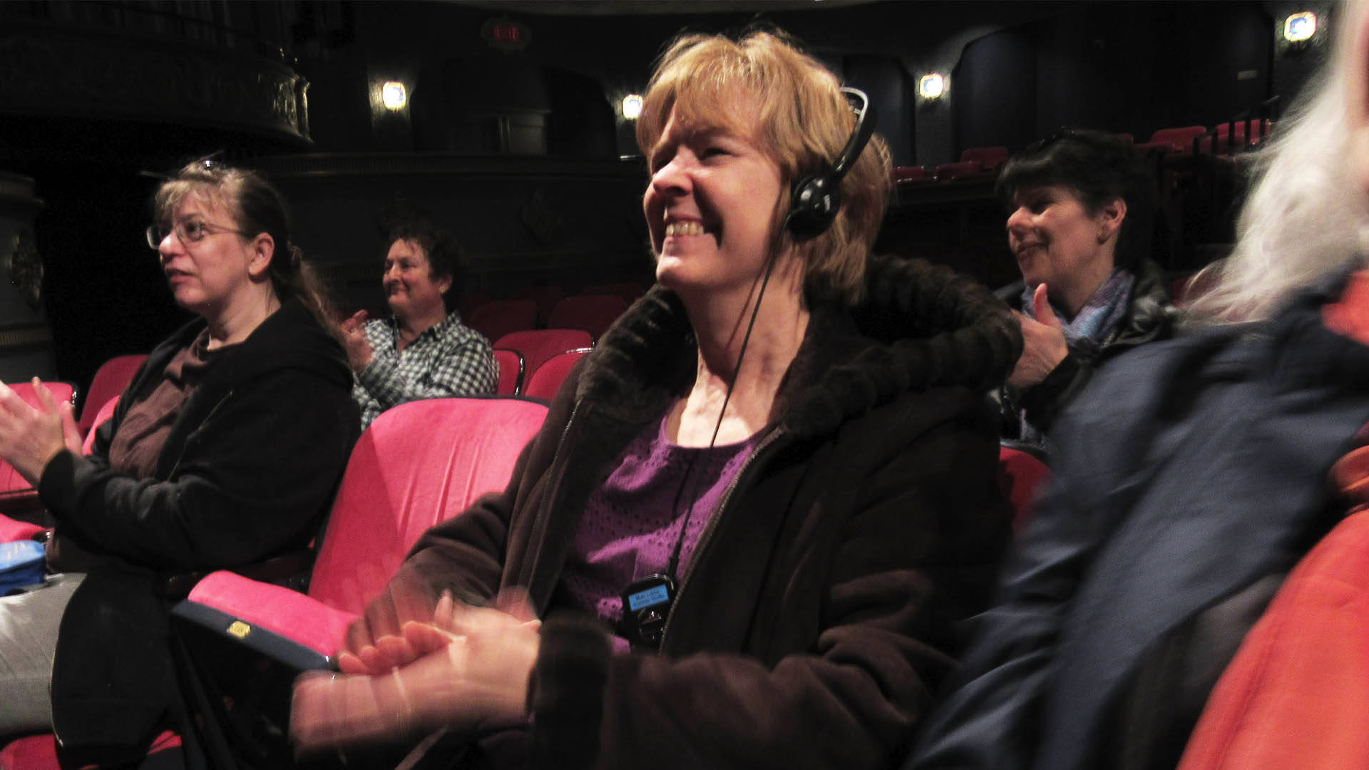 A white woman with strawberry blonde hair sits in a seat with an assisted listening device on.