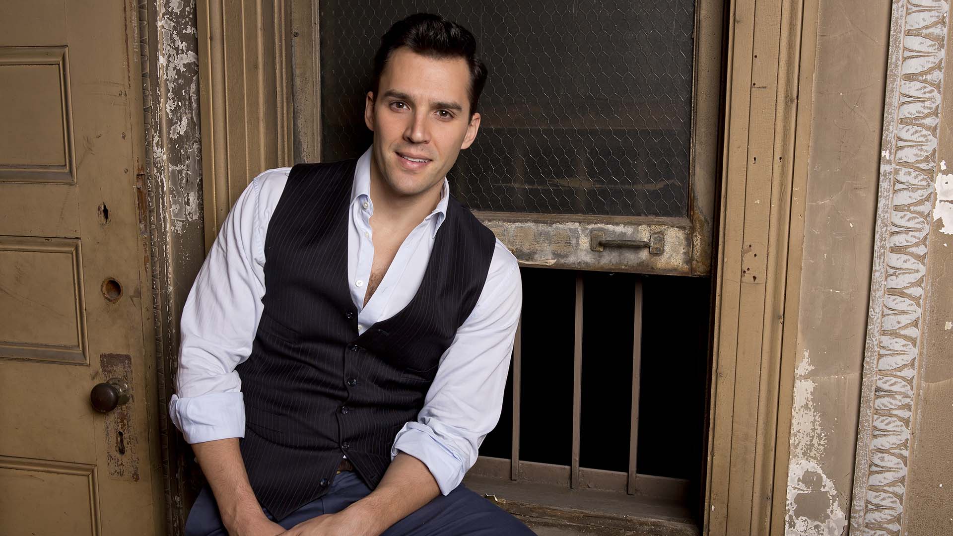 Headshot of Ryan Silverman. He has a handsome youthful face. with short brown hair and brown eyes. He is wearing a buttonup shirt with the top couple buttons open, and a black vest. He is sitting next to an old window with bars and a door.