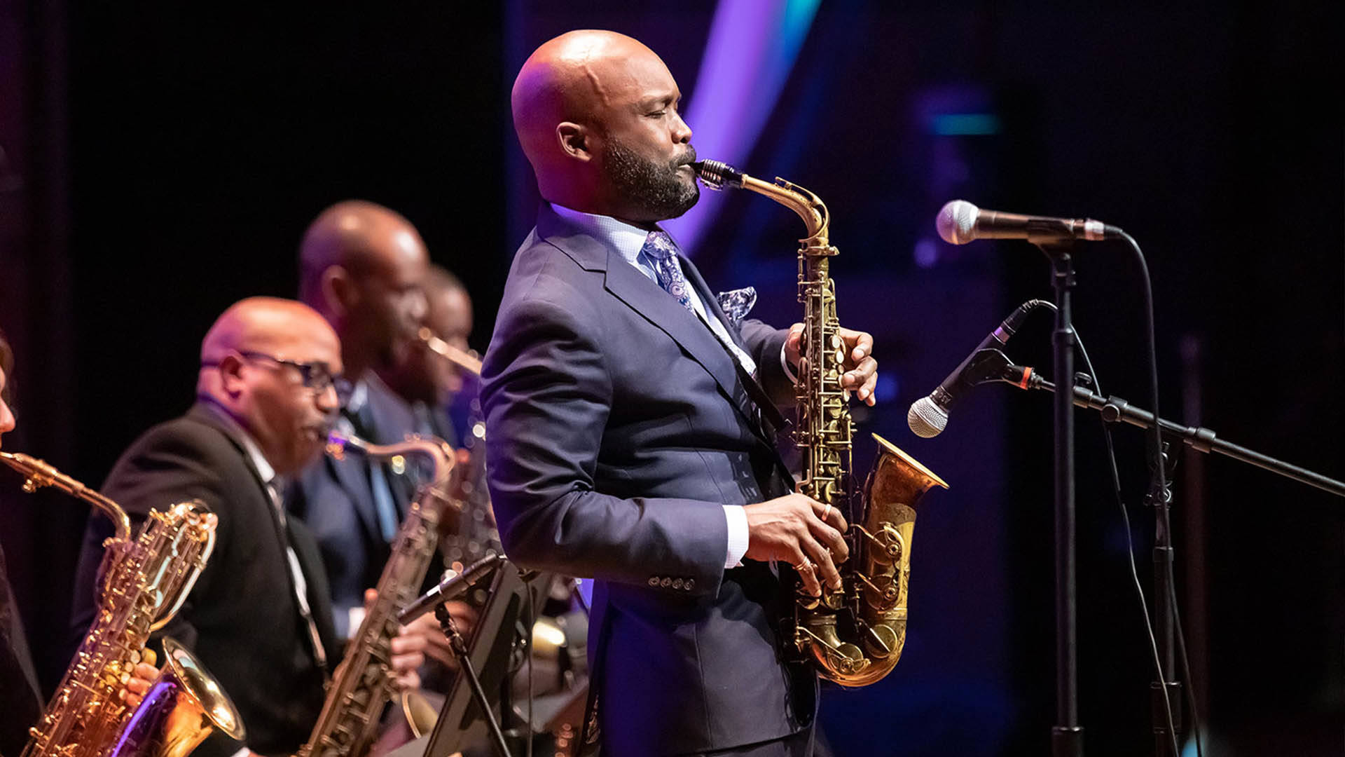 A Black man in a blue suit playing an alto saxophone. 