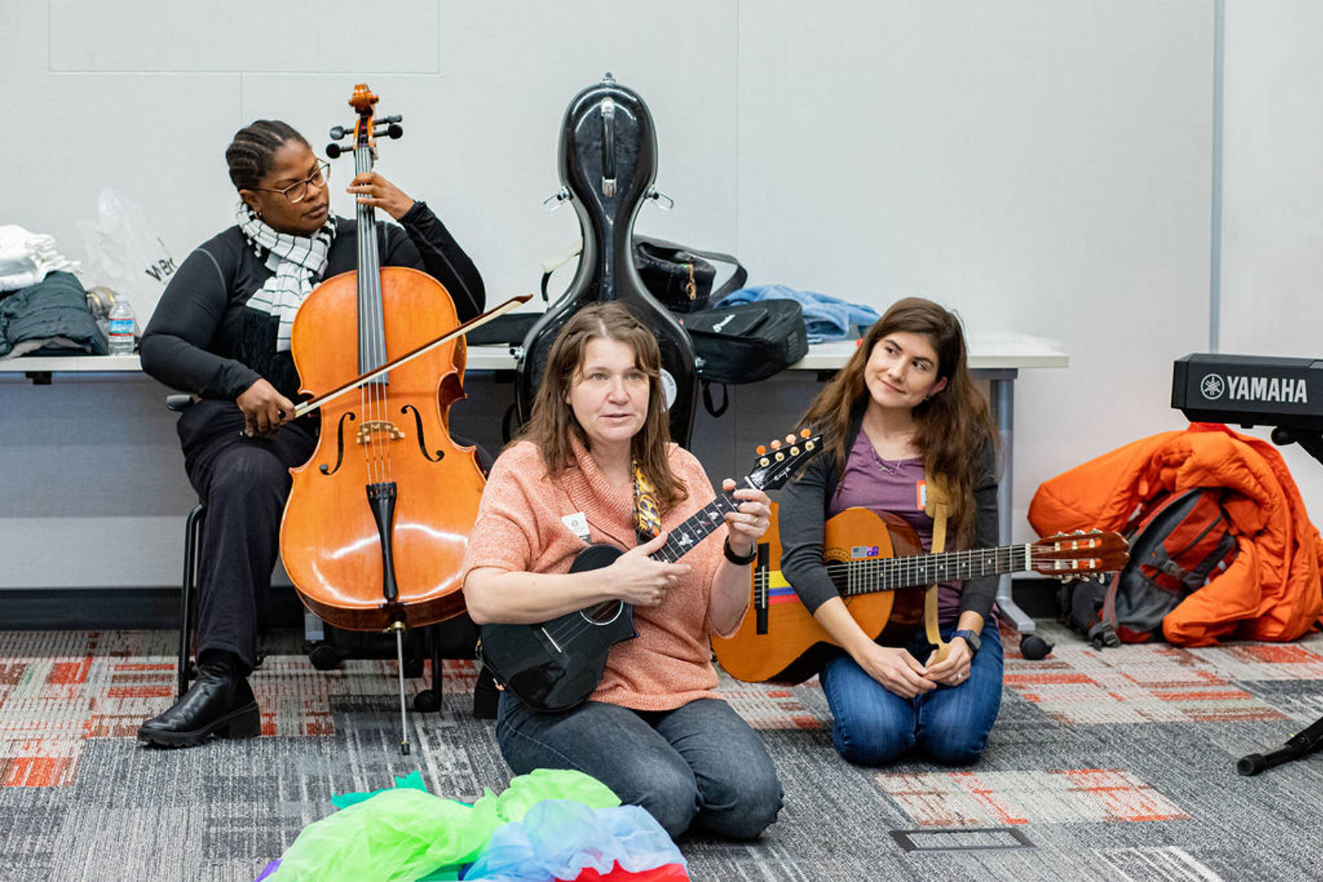 Three people sitting on a carpeted floor holding a cello, a ukelele, and a classical guitar.