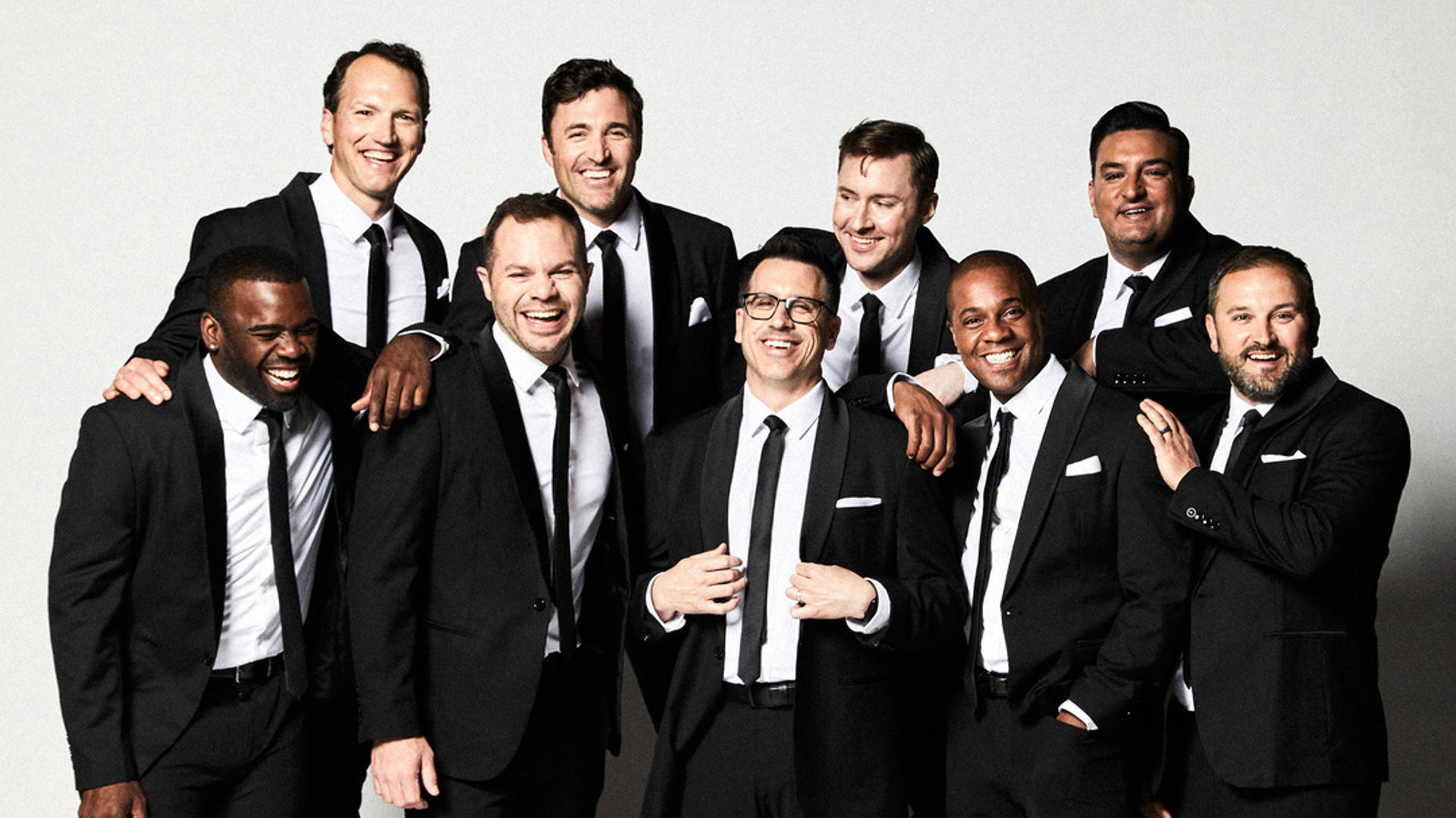 Nine men stand together in two rows posing for a photo. They are all wearing black suits with black ties. They look friendly and happy.