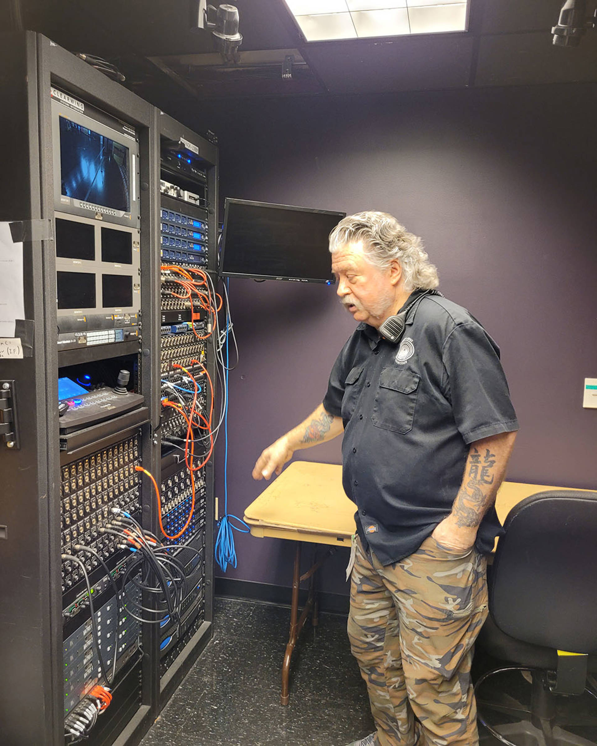 A middle-aged caucasian man with medium length grey hair and a moustache in a black button-down shirt and camoflage pants next to a large switch board with screens and colorful wires.