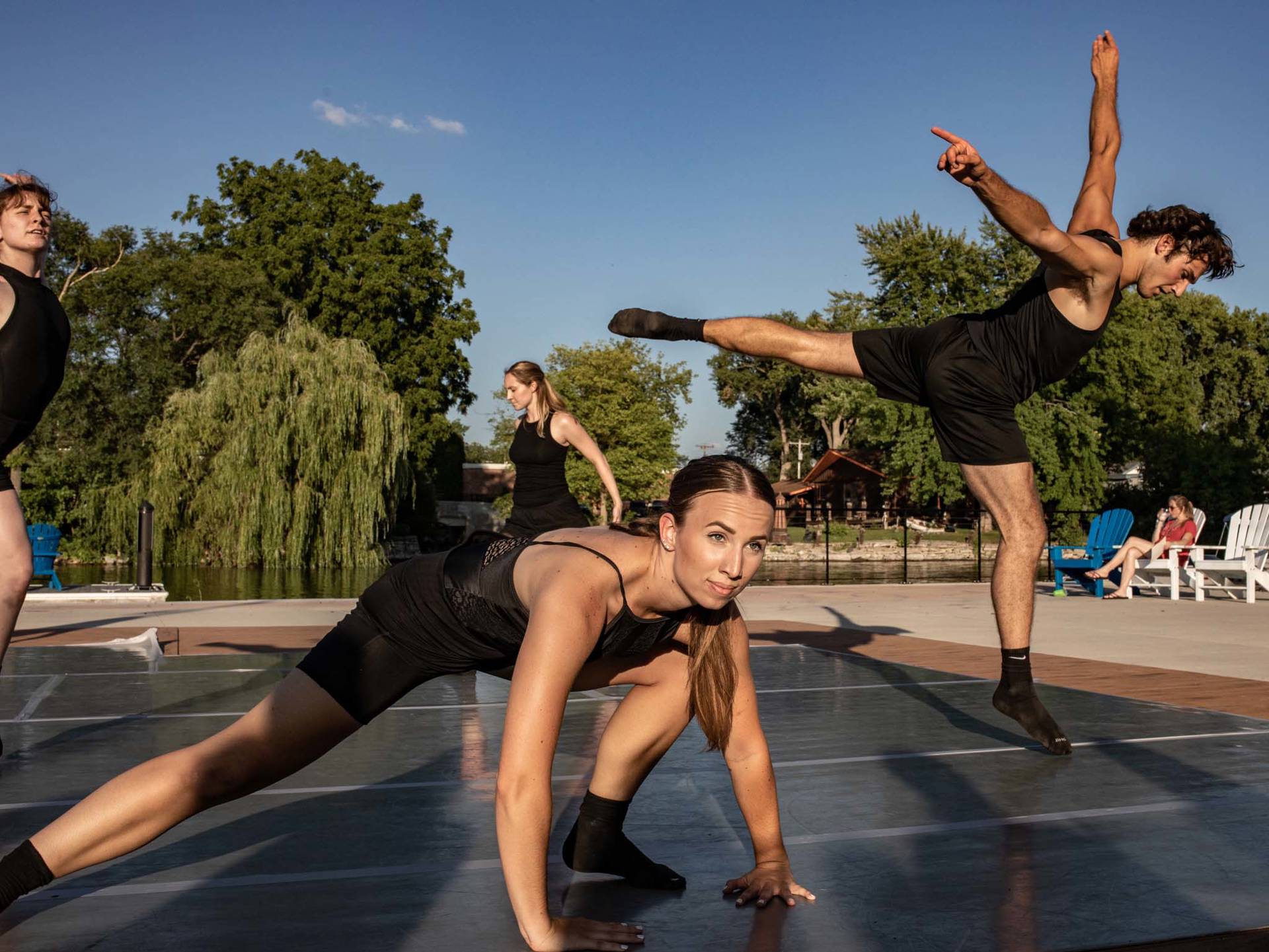 A dancer crouching on her arms and legs. She is on an outdoor stage. Three dancers are in various positions behind her. They are all wearing black.