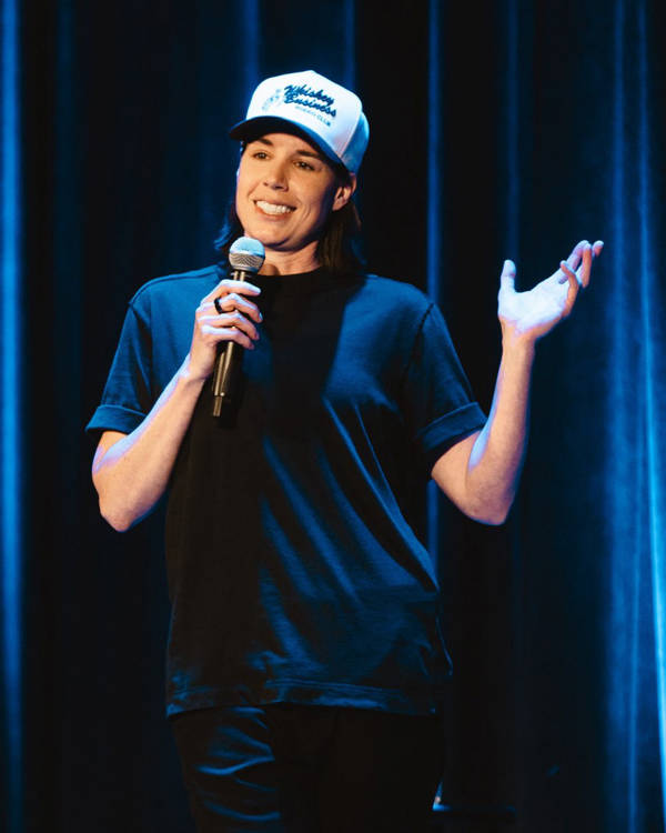 Danae Hays, a woman with light complexion, medium length brown hair, a trucker cap and a black shirt performing standup comedy.