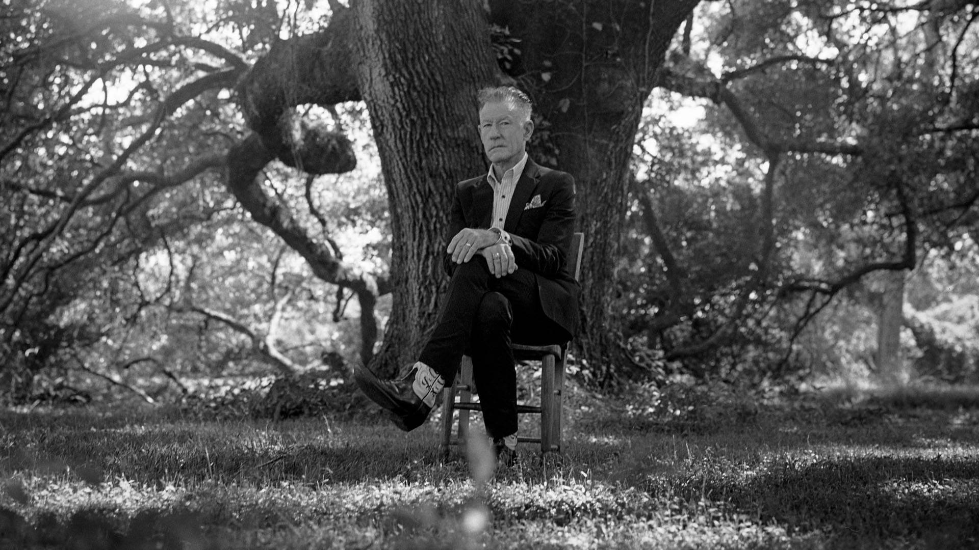 Grayscale photo of an older man with fair skin and short hair sitting on a chair in a suit and cowboy boots. He is outside in front of a large tree and his legs are crossed. He is resting his hands on his knee.
