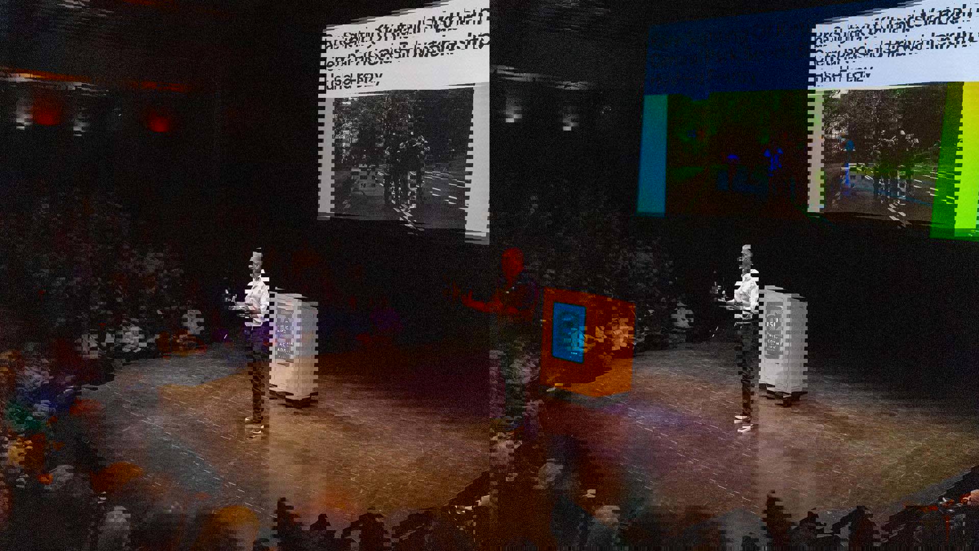 An african american man with buzzed greyish hair, glasses, and a white button-up shirt speaking to a seated audience.