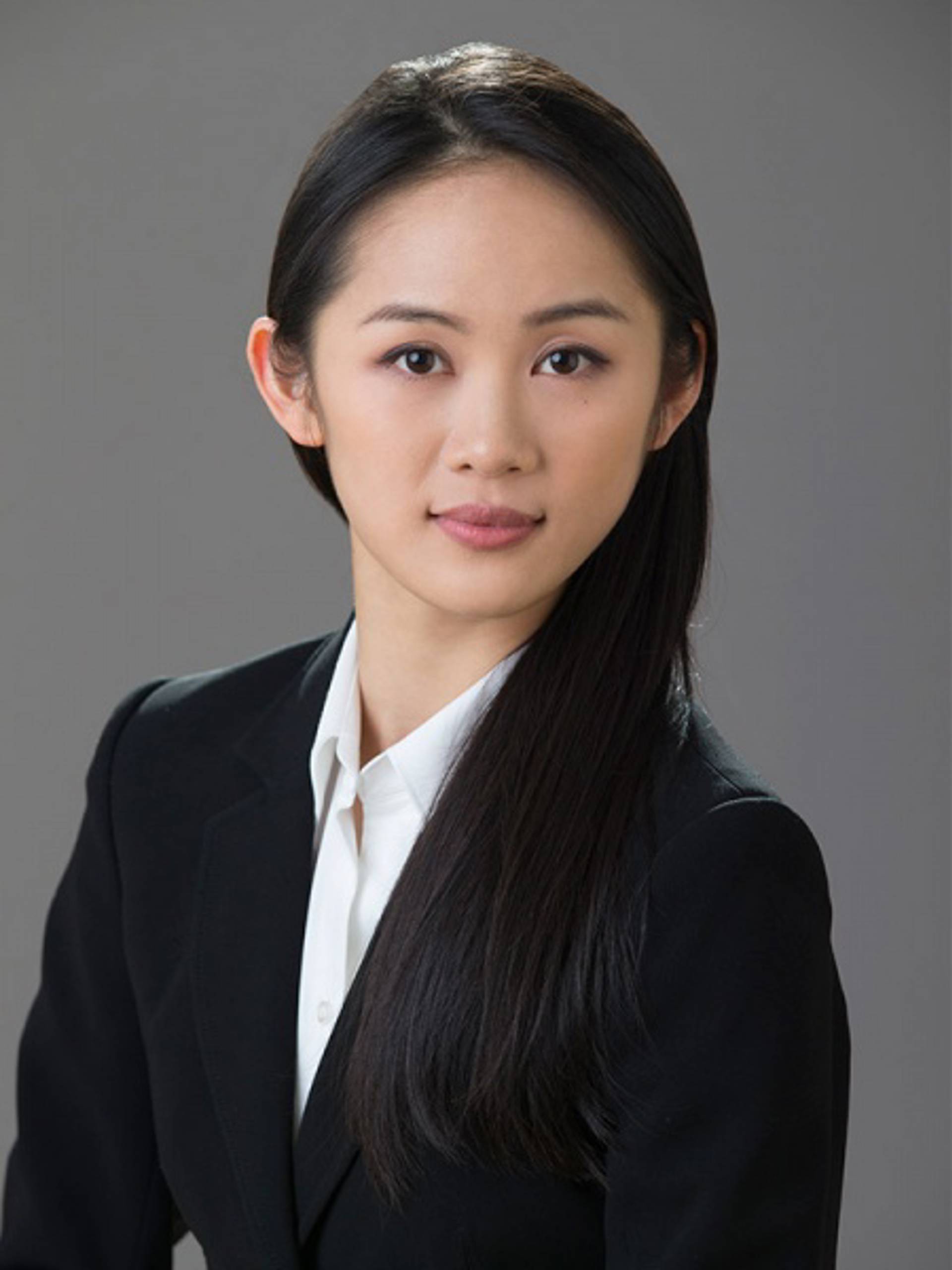 Headshot of a young woman with medium complexion, long black hair, a white collared shirt and a dark jacket against a grey background