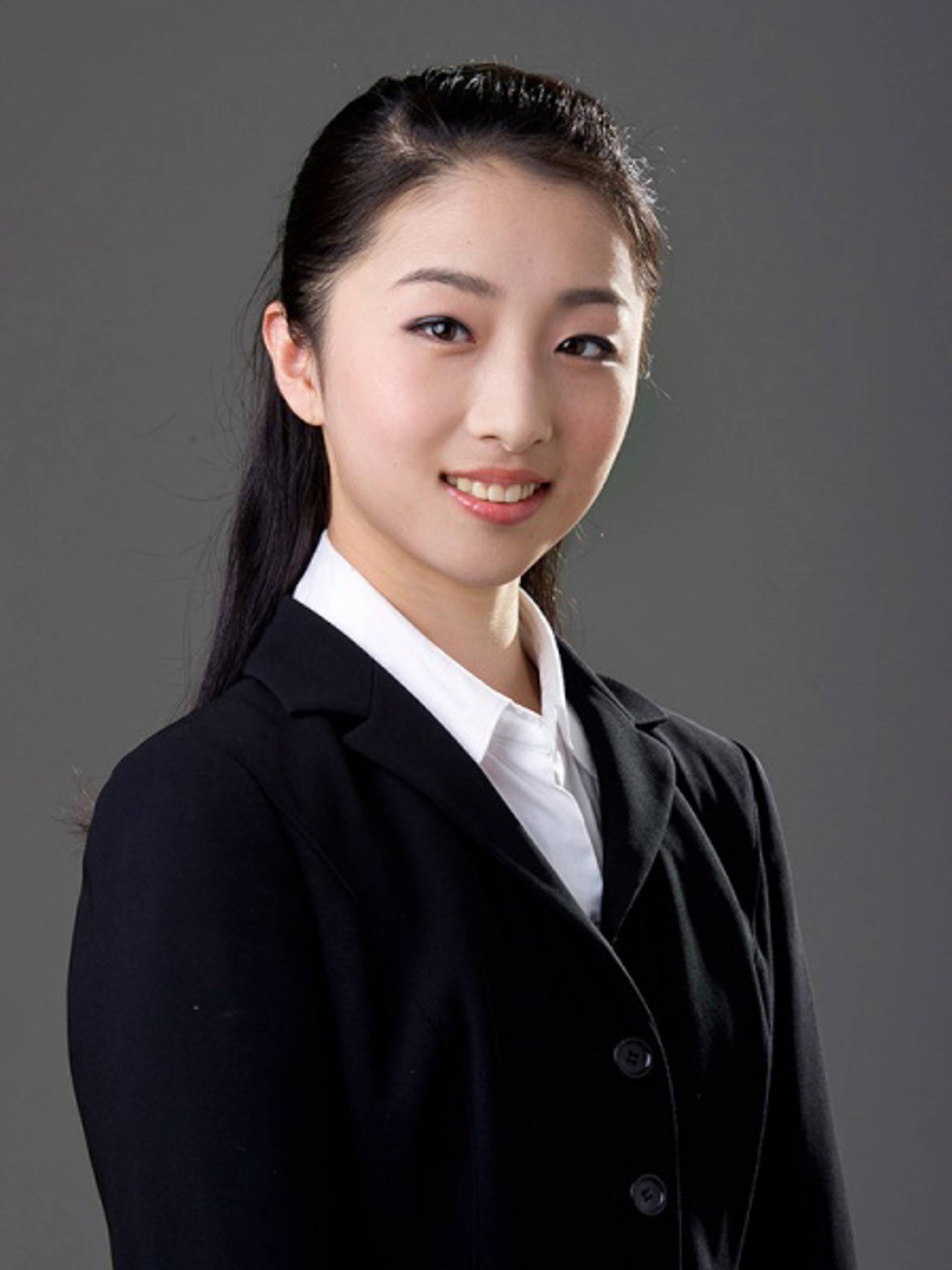 Headshot of a young woman with medium complexion, long black hair, a white collared shirt and a dark jacket against a grey background
