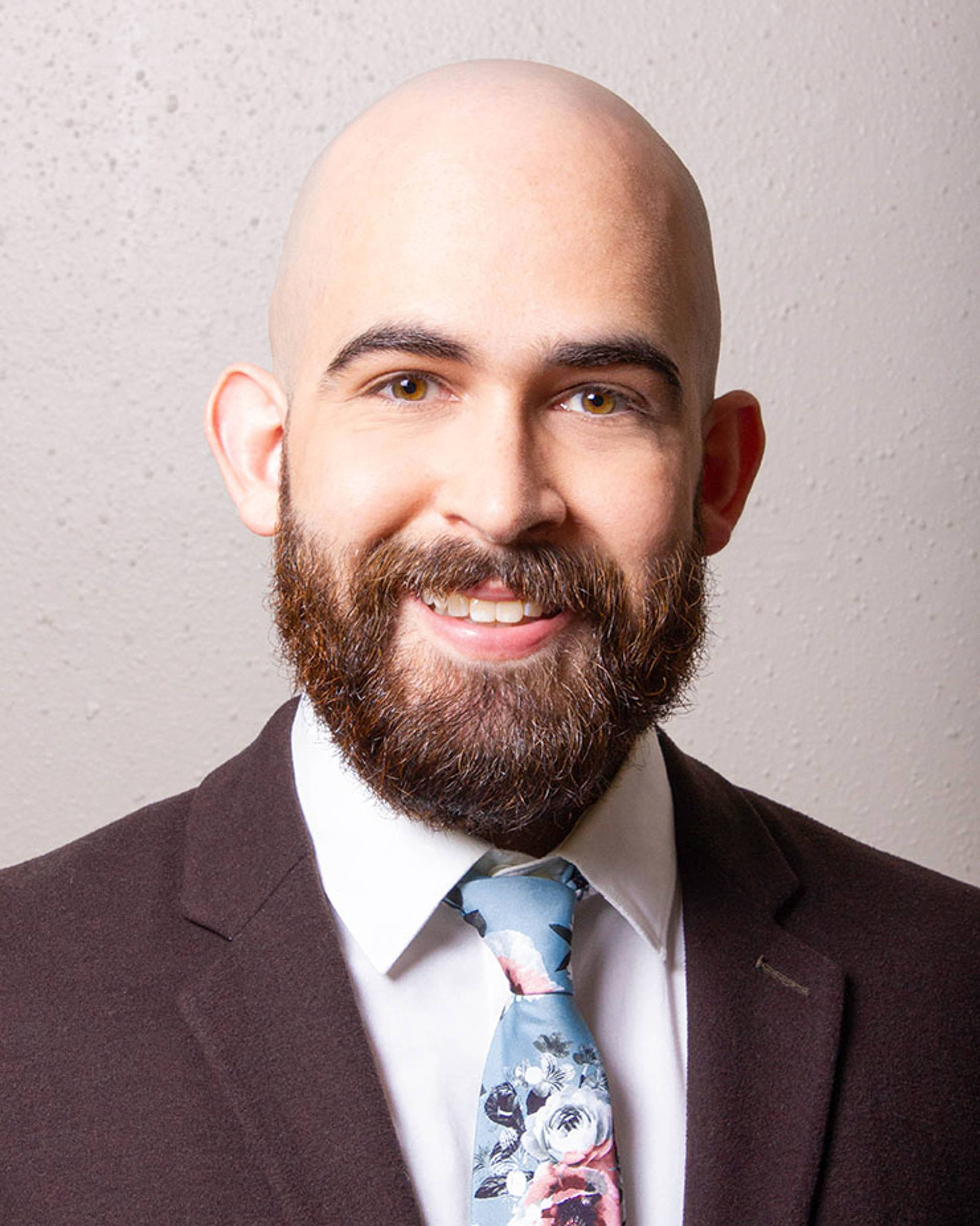 Headshot of a smiling man with brown eyes, a bald head and a full brown beard. He is wearing a dark brown jacket with a floral tie.