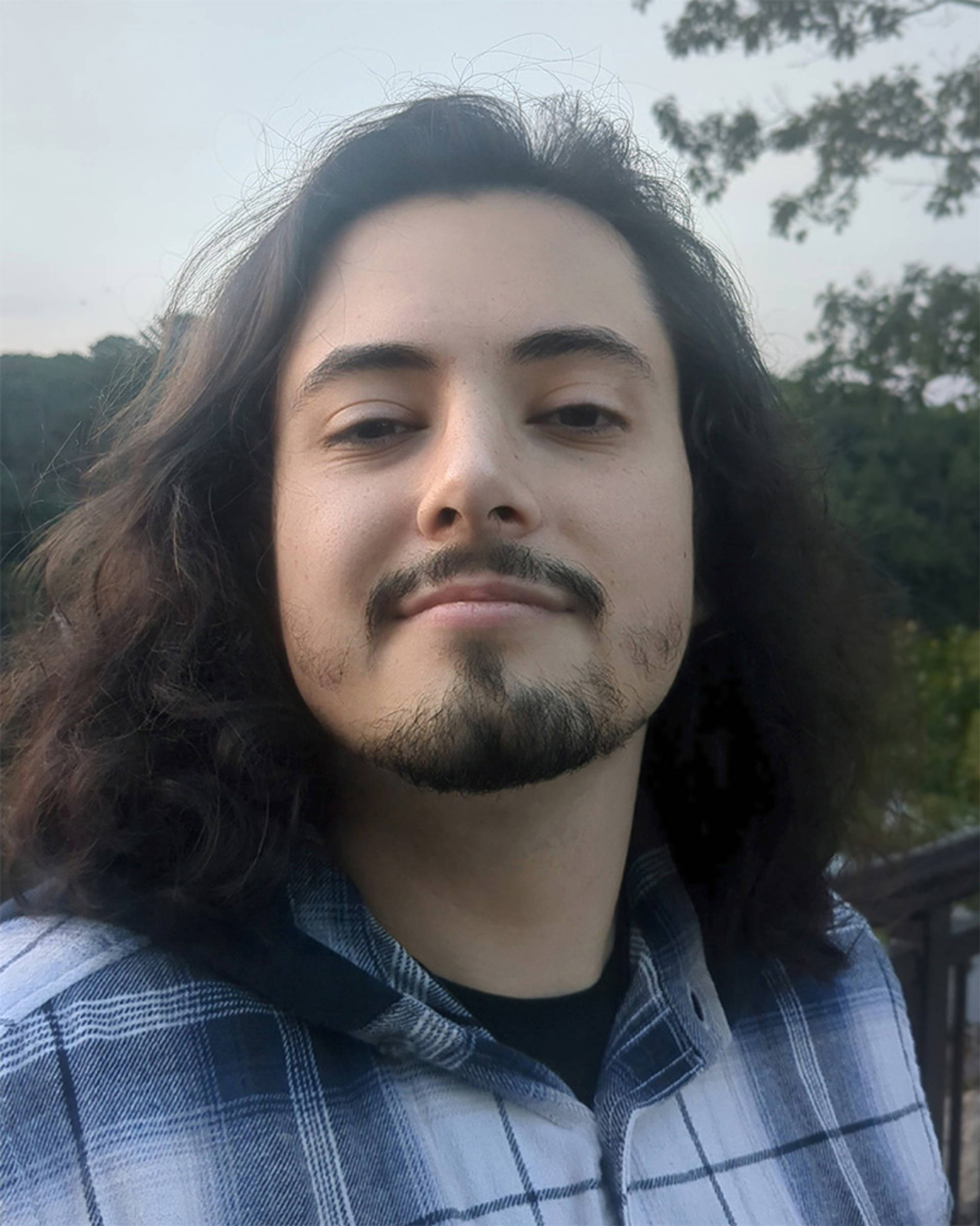 Headshot of a man with long dark hair, dark eyes, facial hair, and a plaid shirt.