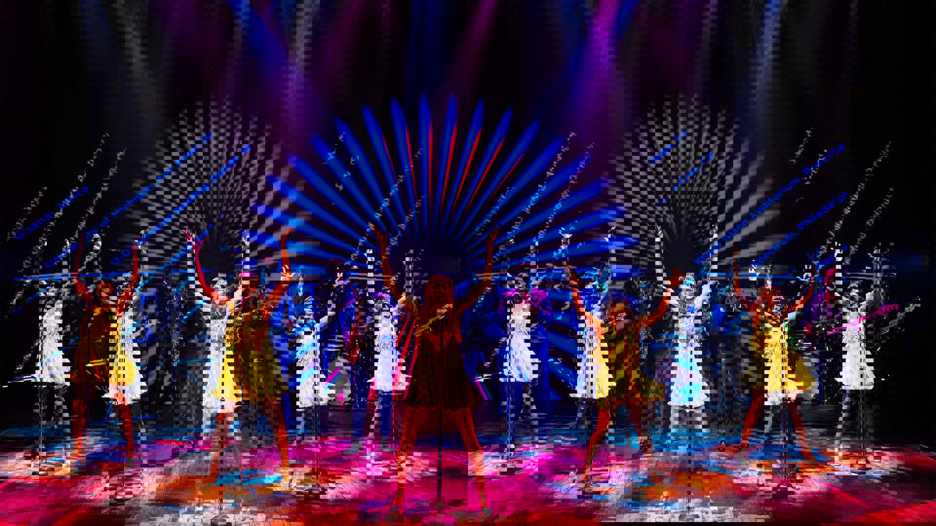 Five black women in short yellow dresses singing into mic stands. A jazz band performs behind them with blue and pink lighting. The background has city names projected with a blue spiral design.