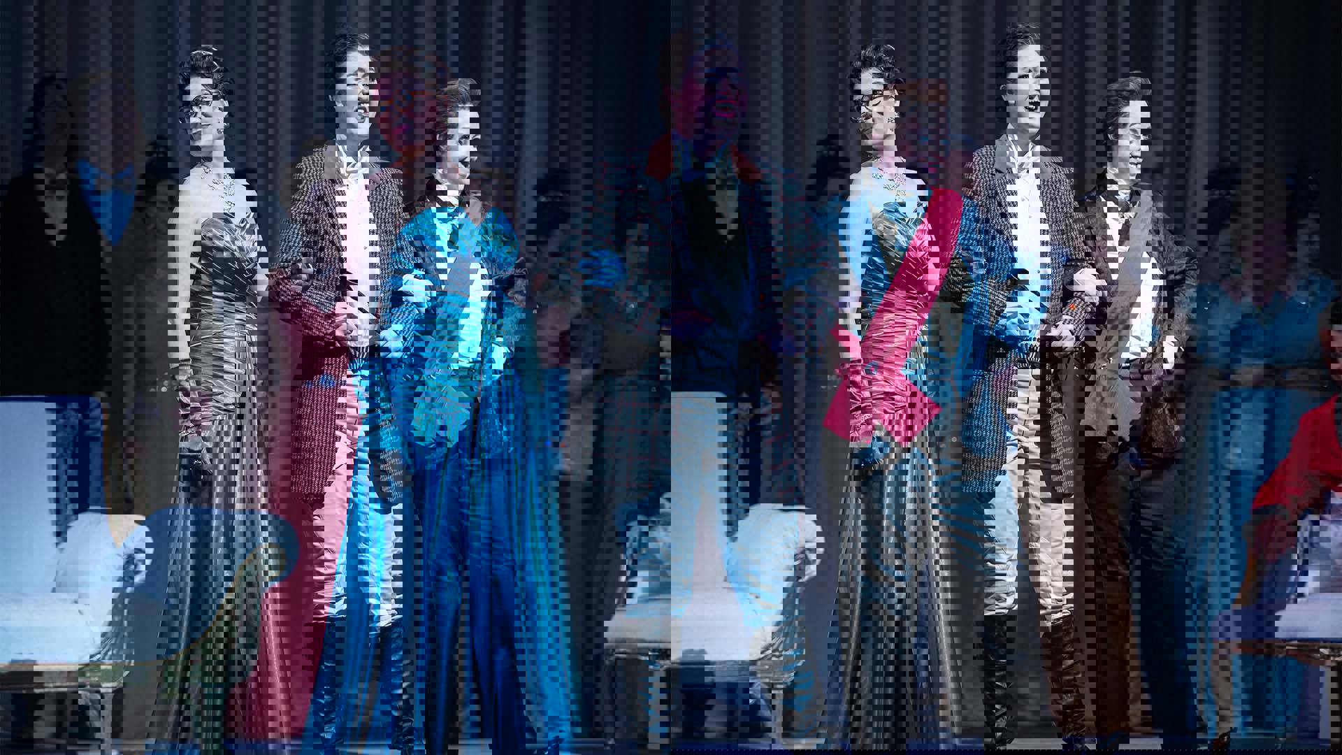 Three caucasian women performing in an opera. They are hooking arms. the center woman has a jockey costume on. the woman on the left has a ballgown on. The woman on the right is wearing a tux with a red sash.